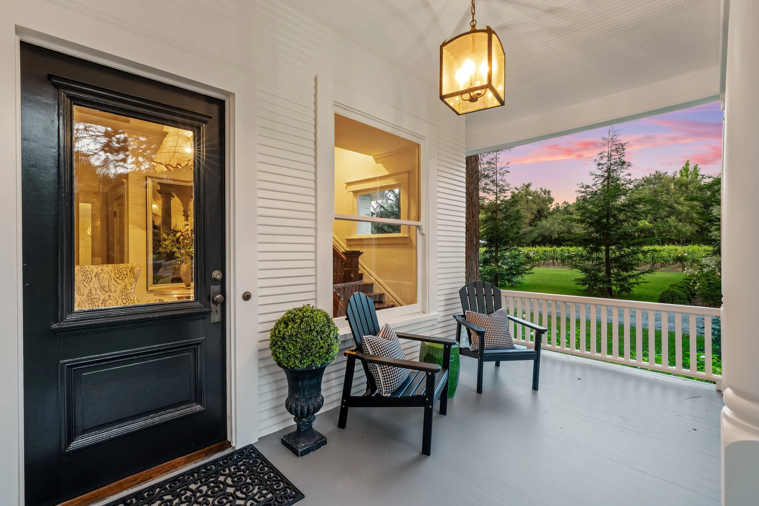Covered porch with white siding, black front door with glass window, hanging lantern light, black chairs with pillows, and potted plant, overlooking trees and sunset sky.