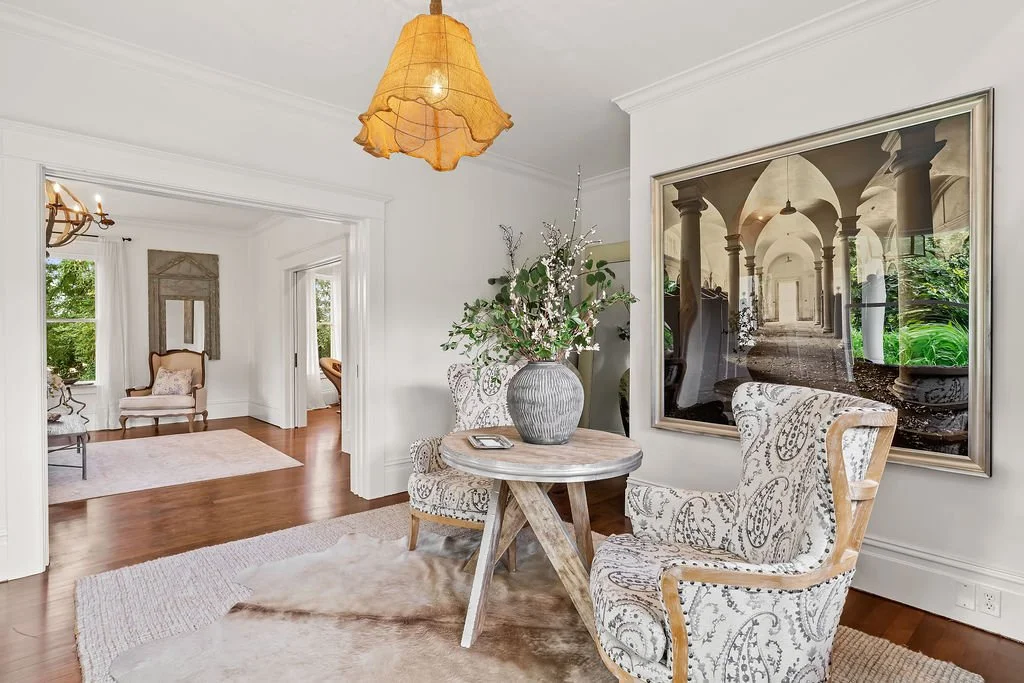 A bright, elegantly decorated living room with white walls, wooden floors, and large windows. There is a circular table with a large vase of flowers, surrounded by patterned armchairs. A large framed photo or painting of an arched corridor is mounted