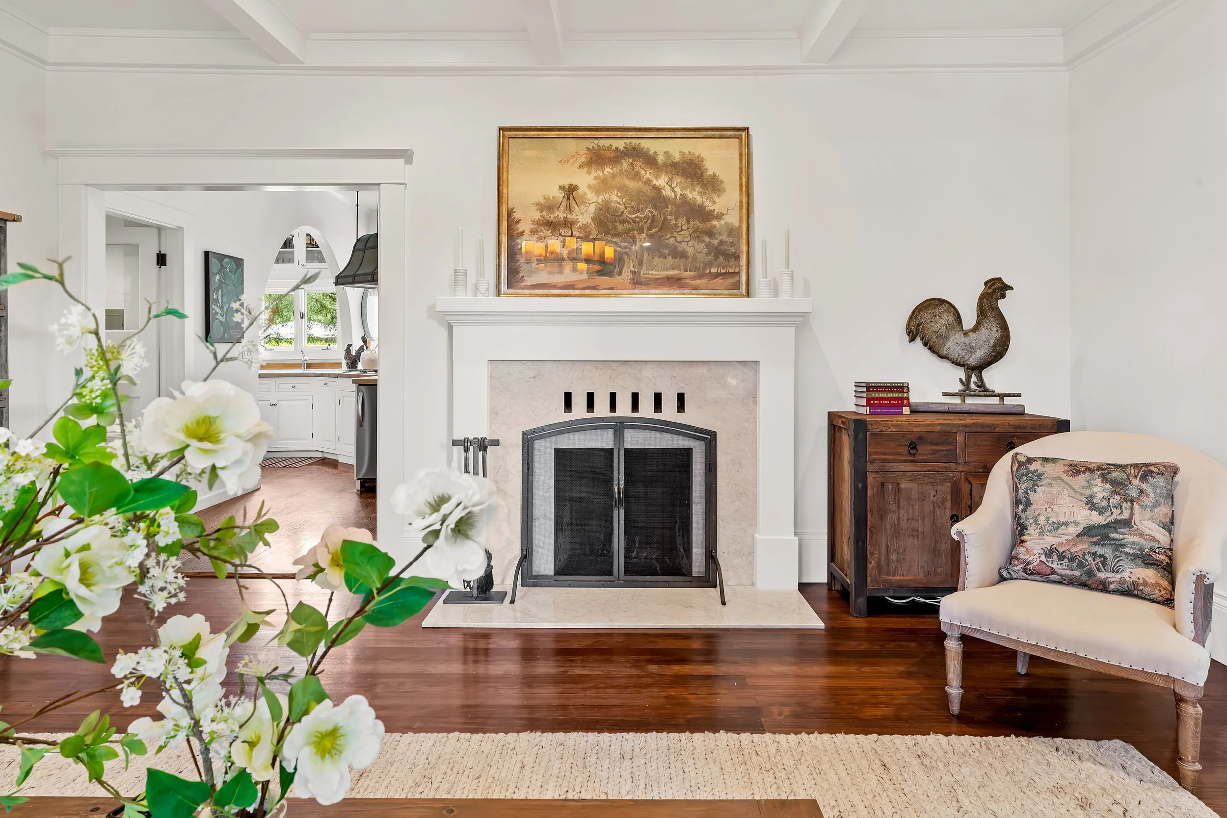 A cozy living room with a fireplace, decorative items, and a floral armchair. A large painting hangs above the fireplace, and a wooden side table with a decorative bird sculpture and books is next to the armchair. The room features white walls, a woo