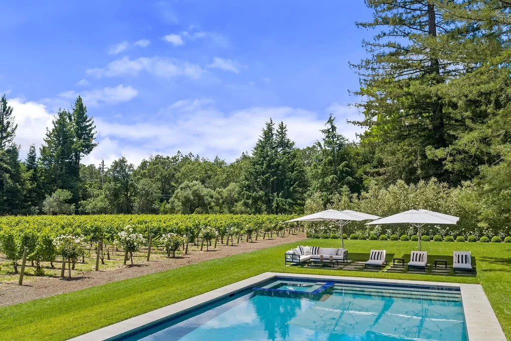 A backyard with a swimming pool, outdoor furniture with umbrellas, green lawn, vineyard in the background, and trees under a partly cloudy sky.
