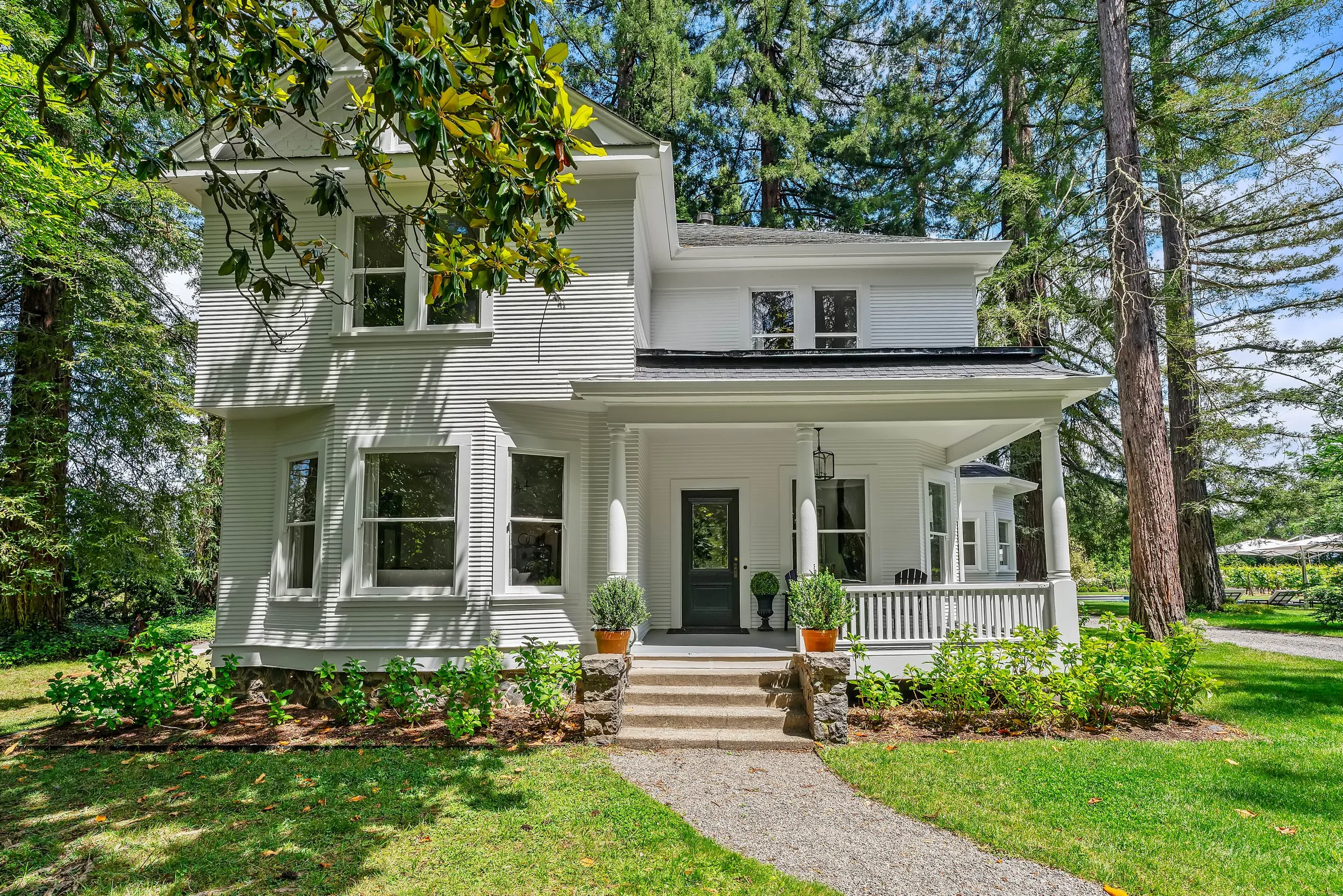 White two-story house with a front porch, surrounded by trees and greenery, with a pathway leading to the entrance.
