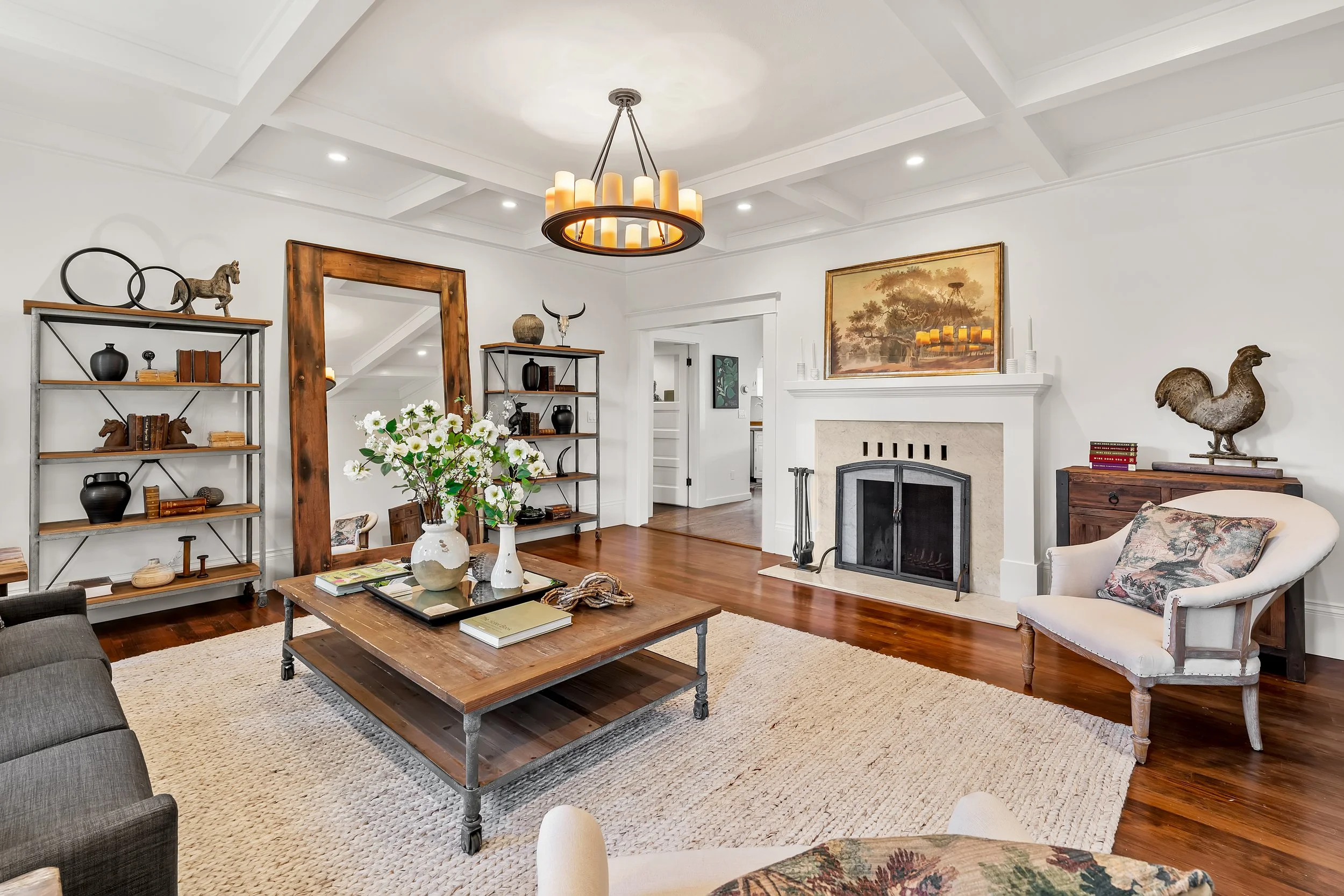 Living room with white walls, hardwood floors, a fireplace, a large wood coffee table with decorative items, two bookshelves with decor, an upholstered armchair, a floral armchair, a gray sofa, and a modern chandelier hanging from a coffered ceiling.