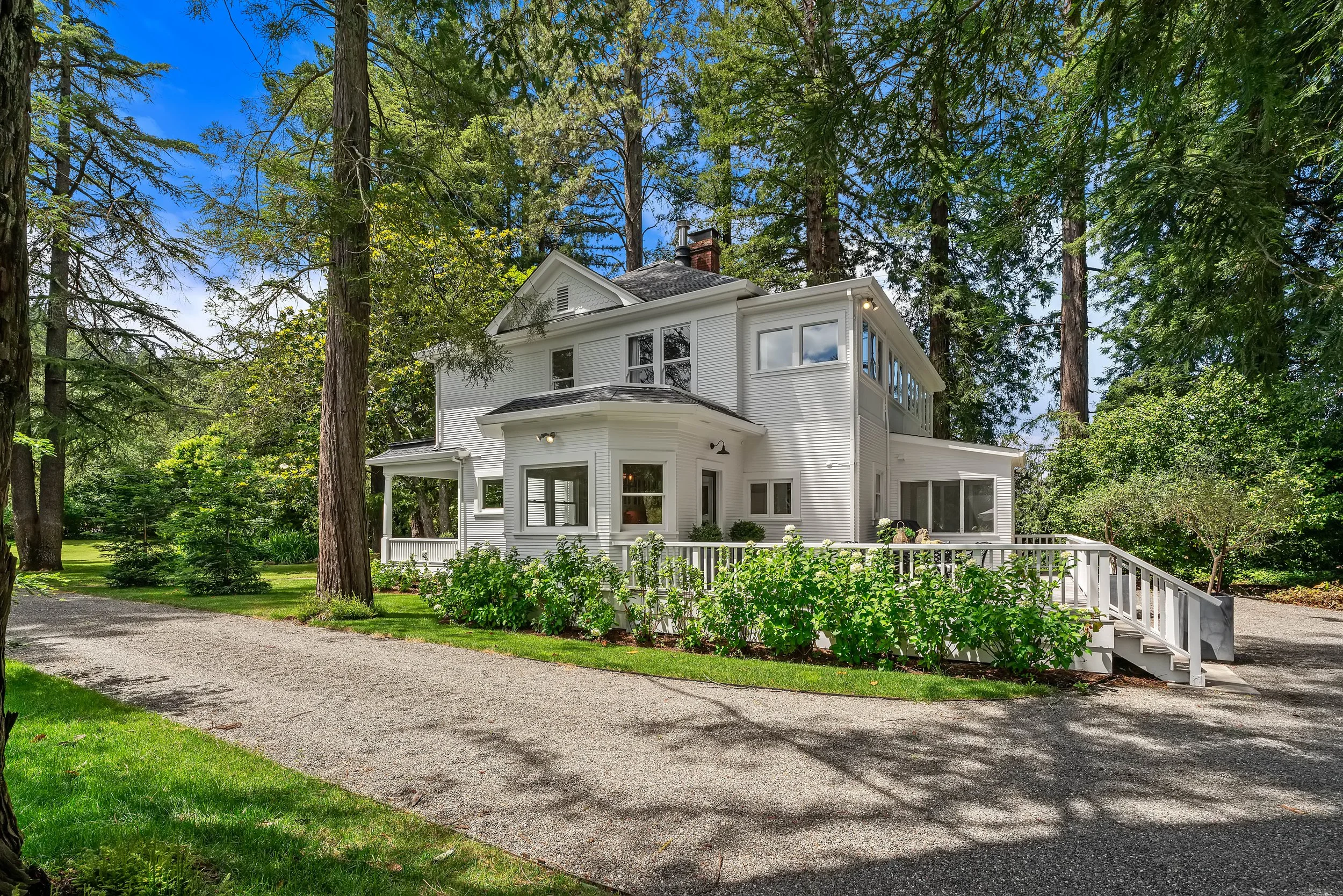 A white two-story house with a black roof, surrounded by tall trees and greenery, with a gravel driveway and a white wooden fence leading to the entrance.