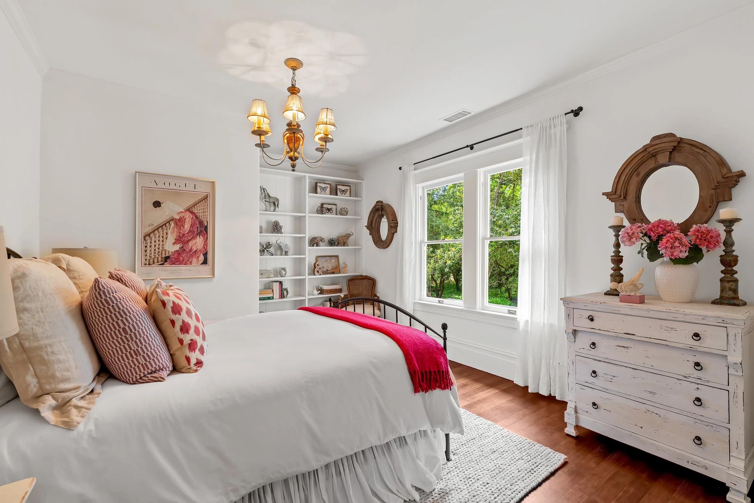 A cozy bedroom with white walls, a bed with white bedding and pink patterned pillows, a white distressed dresser with pink flowers in a vase, a small wooden mirror, a white bookshelf with decorative items and books, large windows with white curtains,