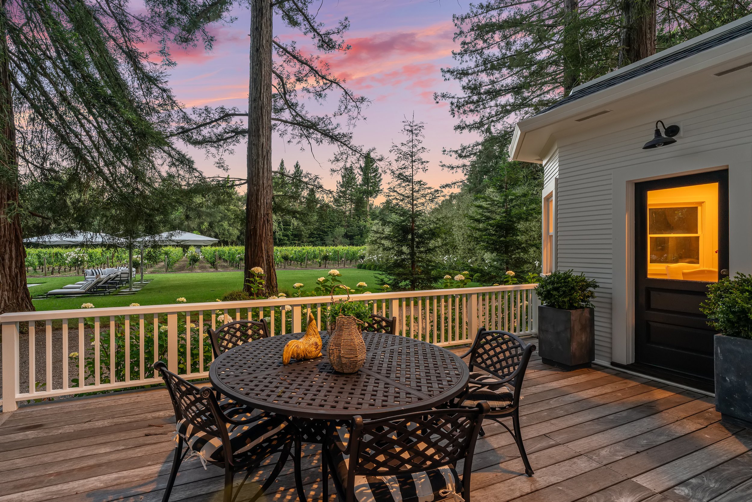 A wooden porch with black metal outdoor furniture including a round table with a small orange cat figurine, a woven basket, and a small potted plant. The porch overlooks a green backyard with tall trees, a vineyard, and patio chairs under umbrellas. 
