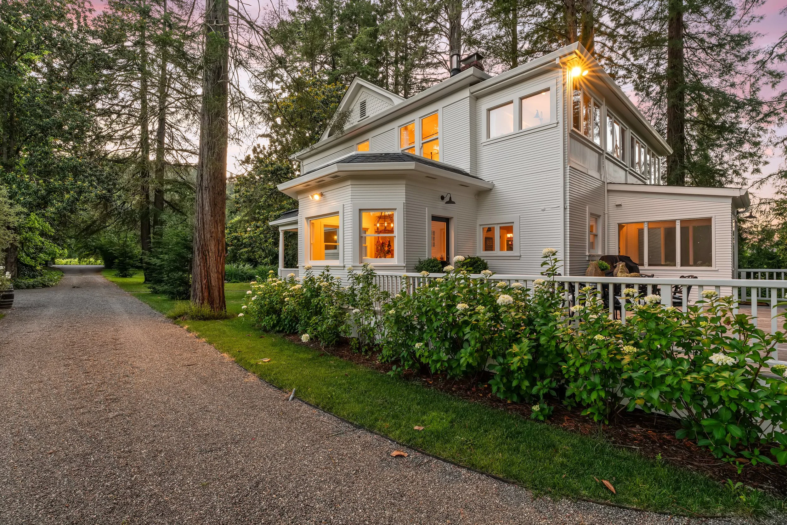 A white multi-story house with large windows illuminated from inside, surrounded by tall trees and bushes, with a gravel driveway leading up to it at dusk.