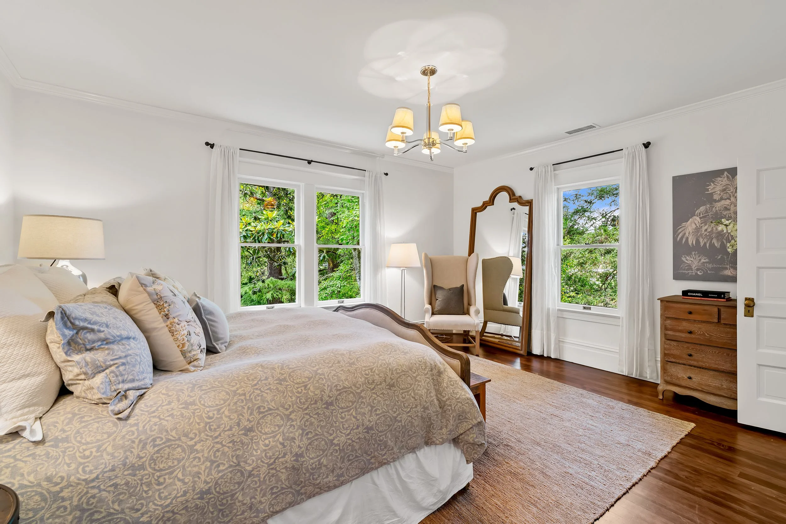 A bedroom with a large bed with beige and blue floral pillows, window view of greenery outside, a chair with a mirror, a nightstand with a lamp, and a wooden dresser, with white walls and curtains.