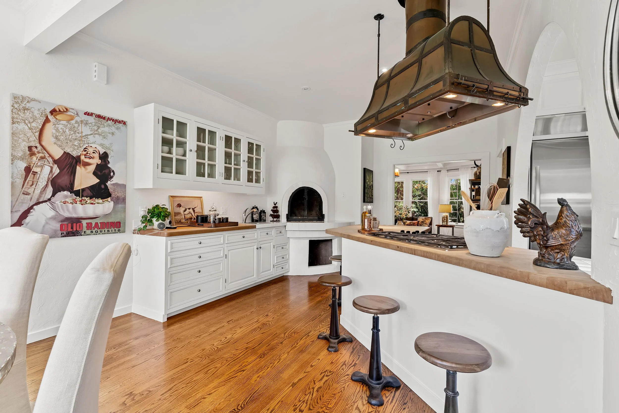 A bright white kitchen with a wooden countertop, barstools, white cabinets with glass doors, and a vintage-style range hood over the stove. There is a large poster on the wall depicting a woman holding a ladle with an image of a bowl of soup, and the