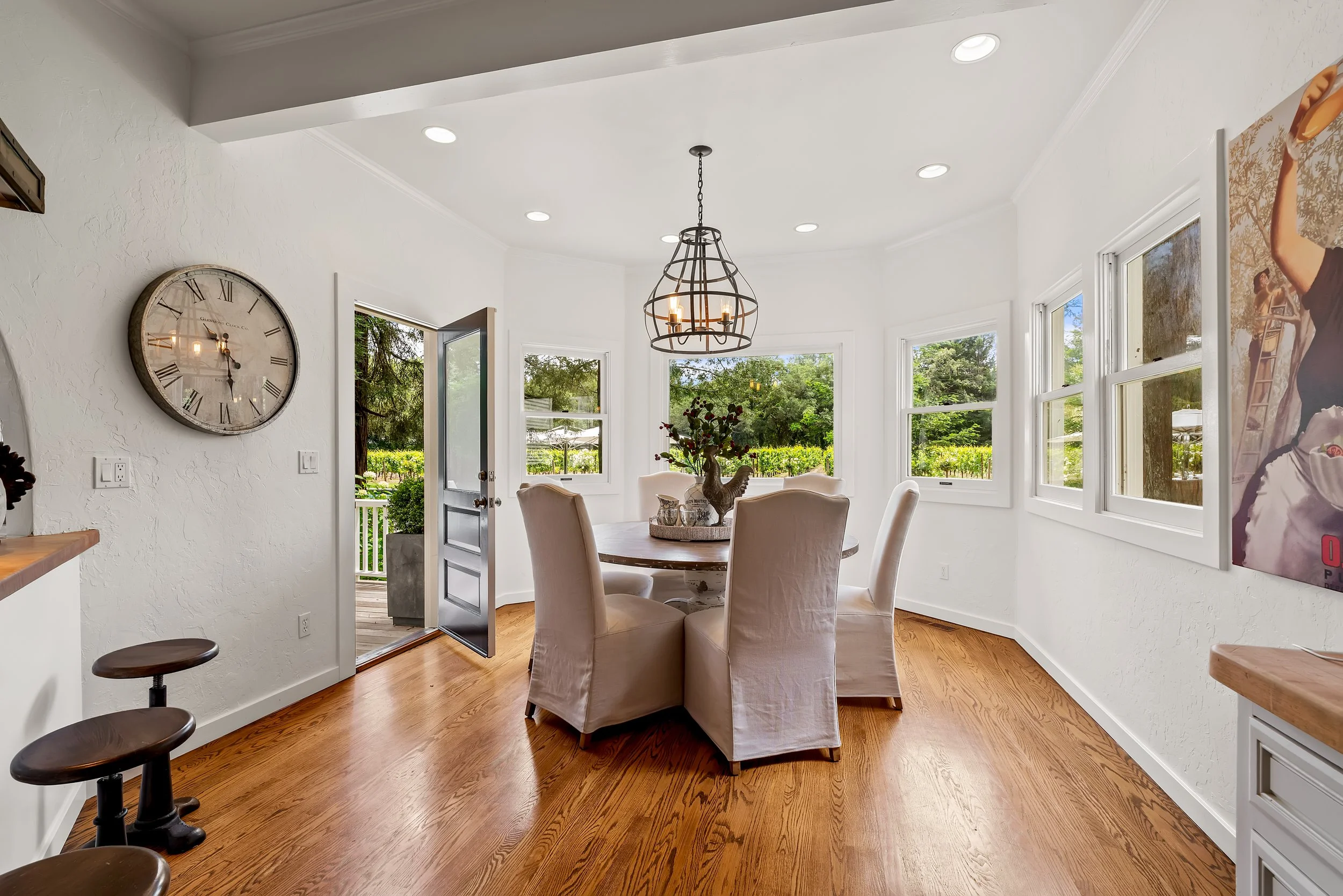 A bright dining area with a round wooden table surrounded by beige upholstered chairs, large windows showing greenery outside, a decorative rooster centerpiece, a wall clock, and a chandelier hanging from the ceiling.