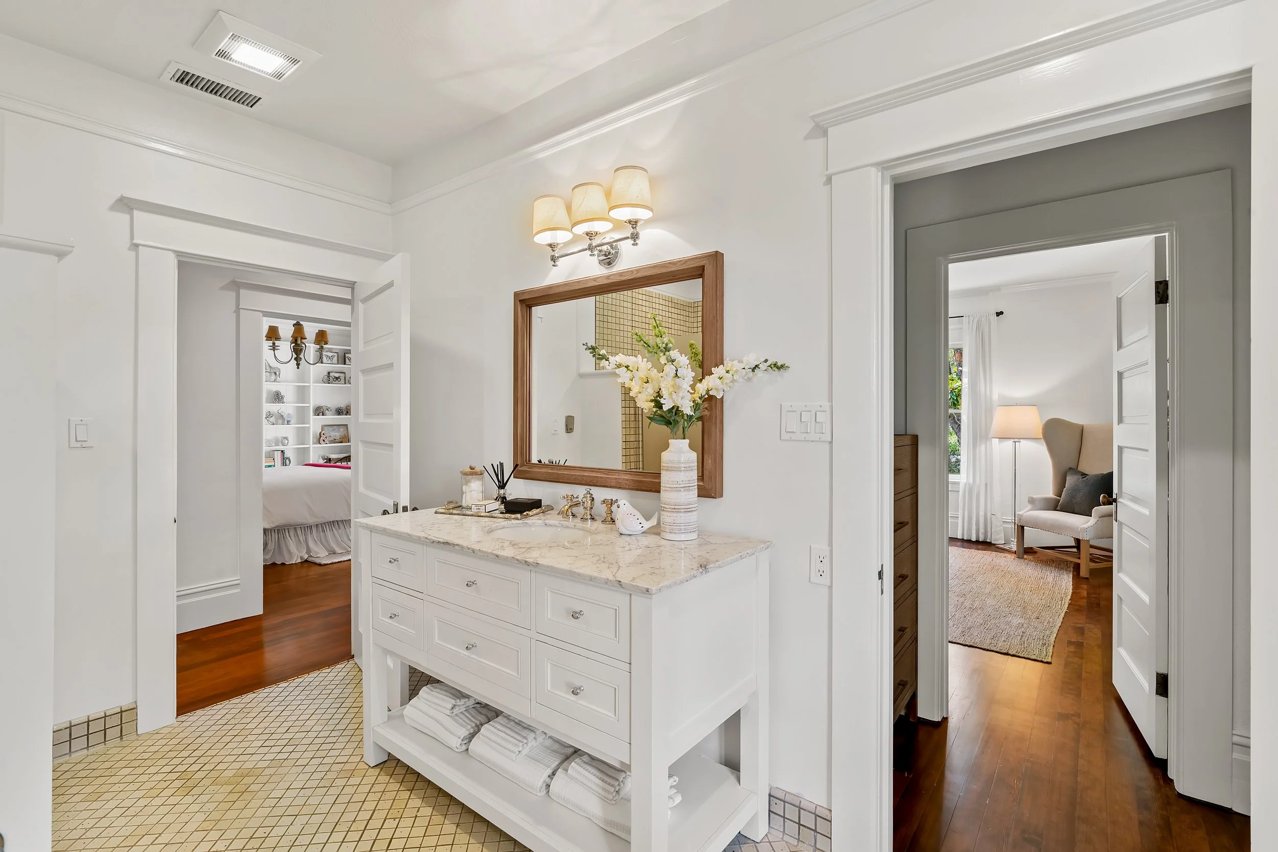 A bathroom area with white walls and cabinetry, a marble countertop with a flower vase, and a mirror. Visible through doorways are a bedroom with white bedding and a room with a beige armchair and side table. The ceiling has a light fixture, and ther