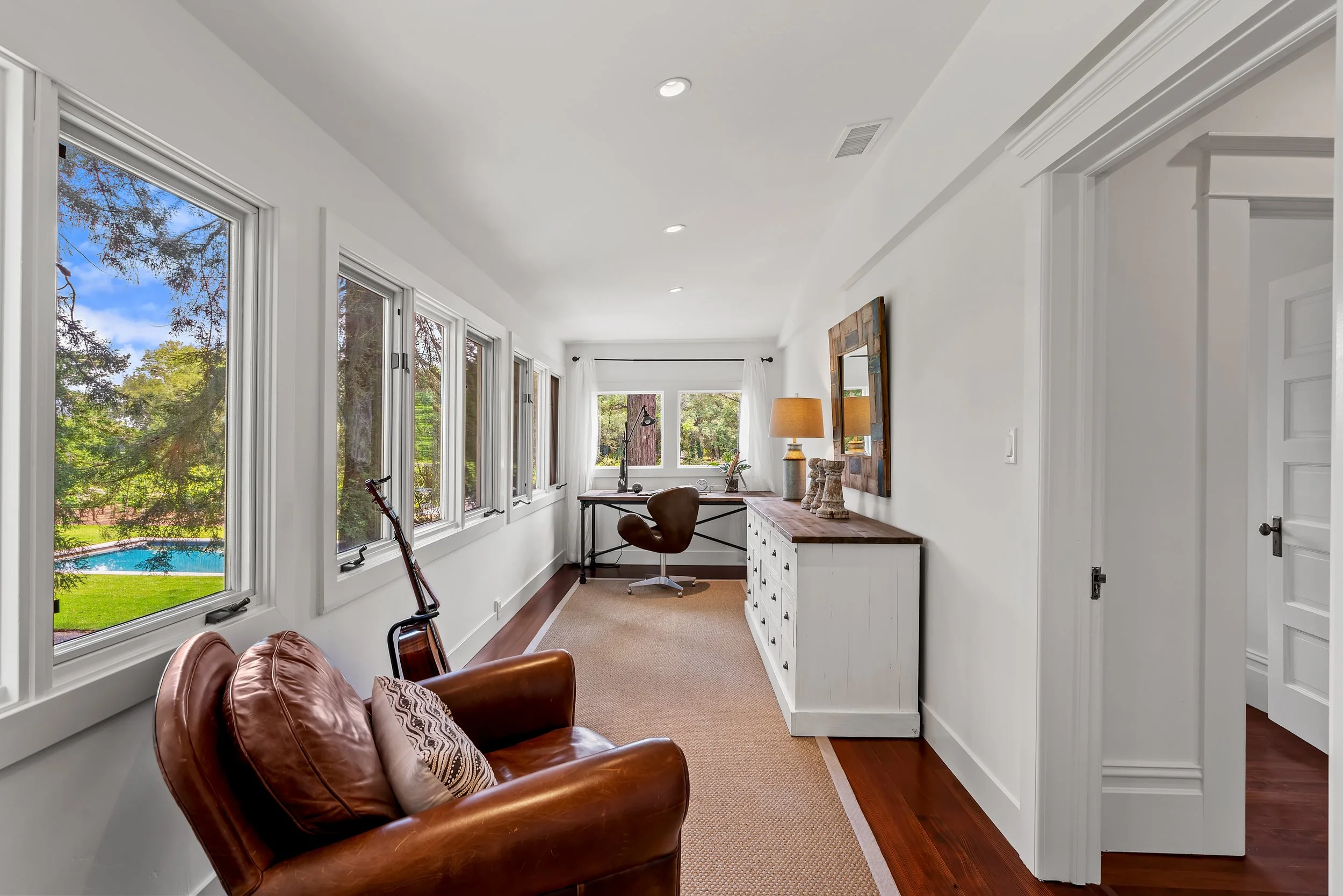 A bright home office with large windows showing trees and a swimming pool outside. The room has a desk, a leather armchair, a lamp, and wall decorations.