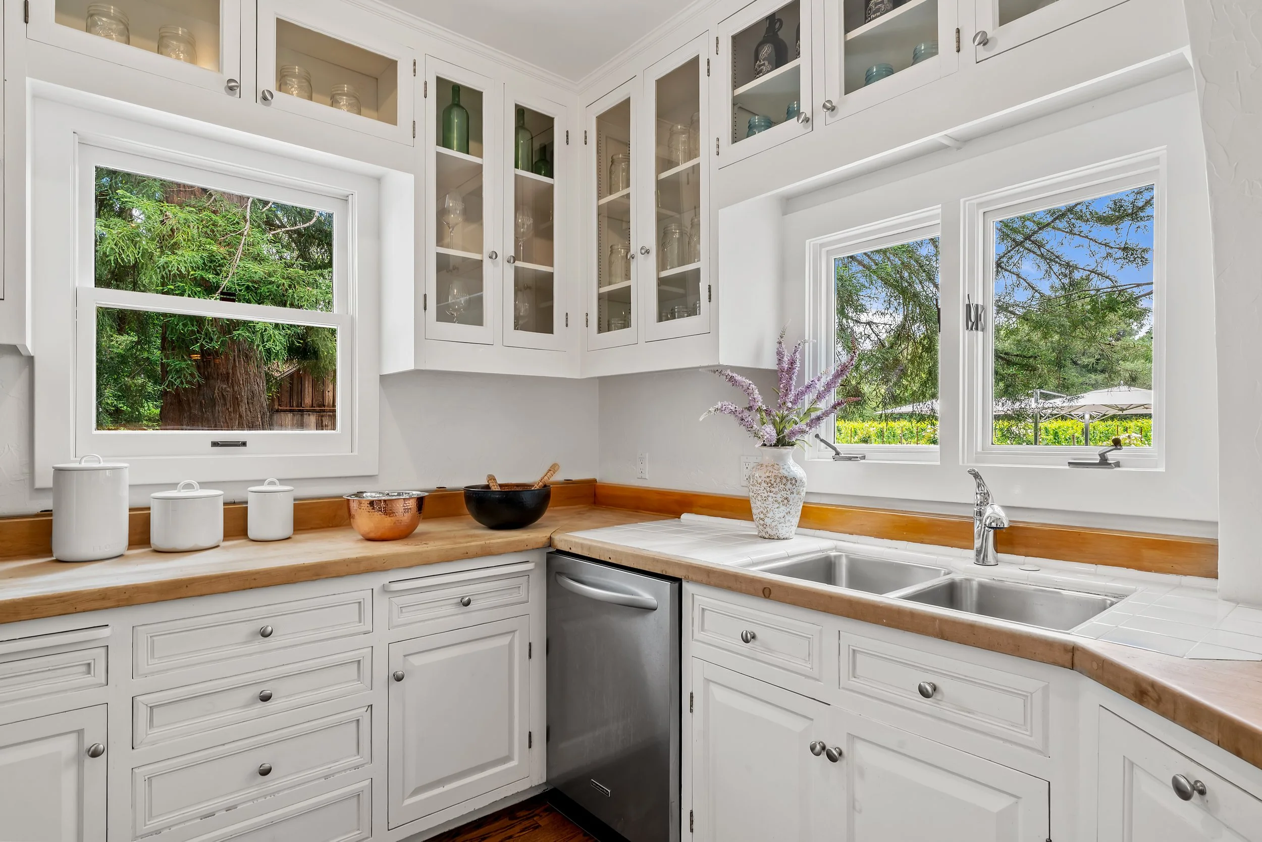 A bright kitchen with white cabinets and multiple glass-front upper cabinets. The kitchen features two large windows overlooking trees and greenery, with a wooden countertop holding various white canisters, a decorative copper bowl, and a black bowl.