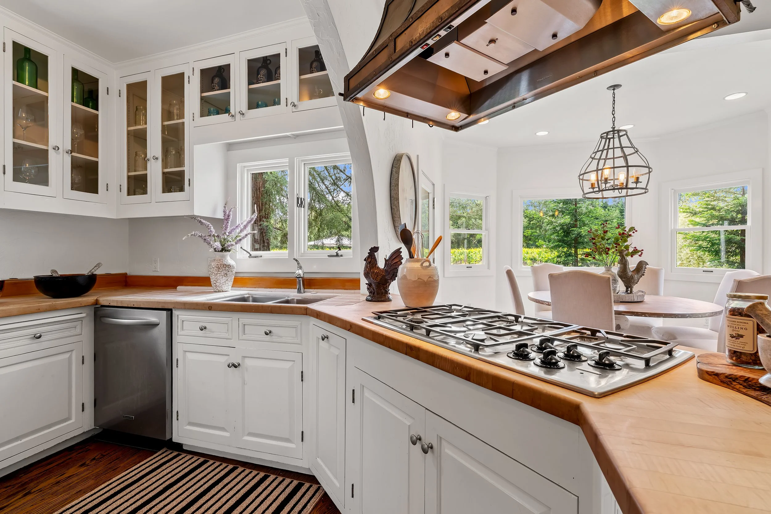 Kitchen with white cabinets, wooden countertops, an island, and large windows showing green trees outside. Decorative vases and figurines are on the counter, and a chandelier hangs over the dining table with chairs.
