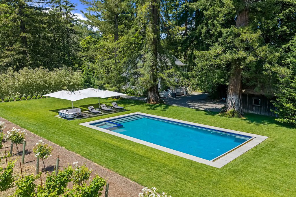 A backyard with a rectangular swimming pool, surrounded by lush green grass, with four lounge chairs under a large white umbrella, large trees providing shade, and some flowering bushes along a gravel pathway.