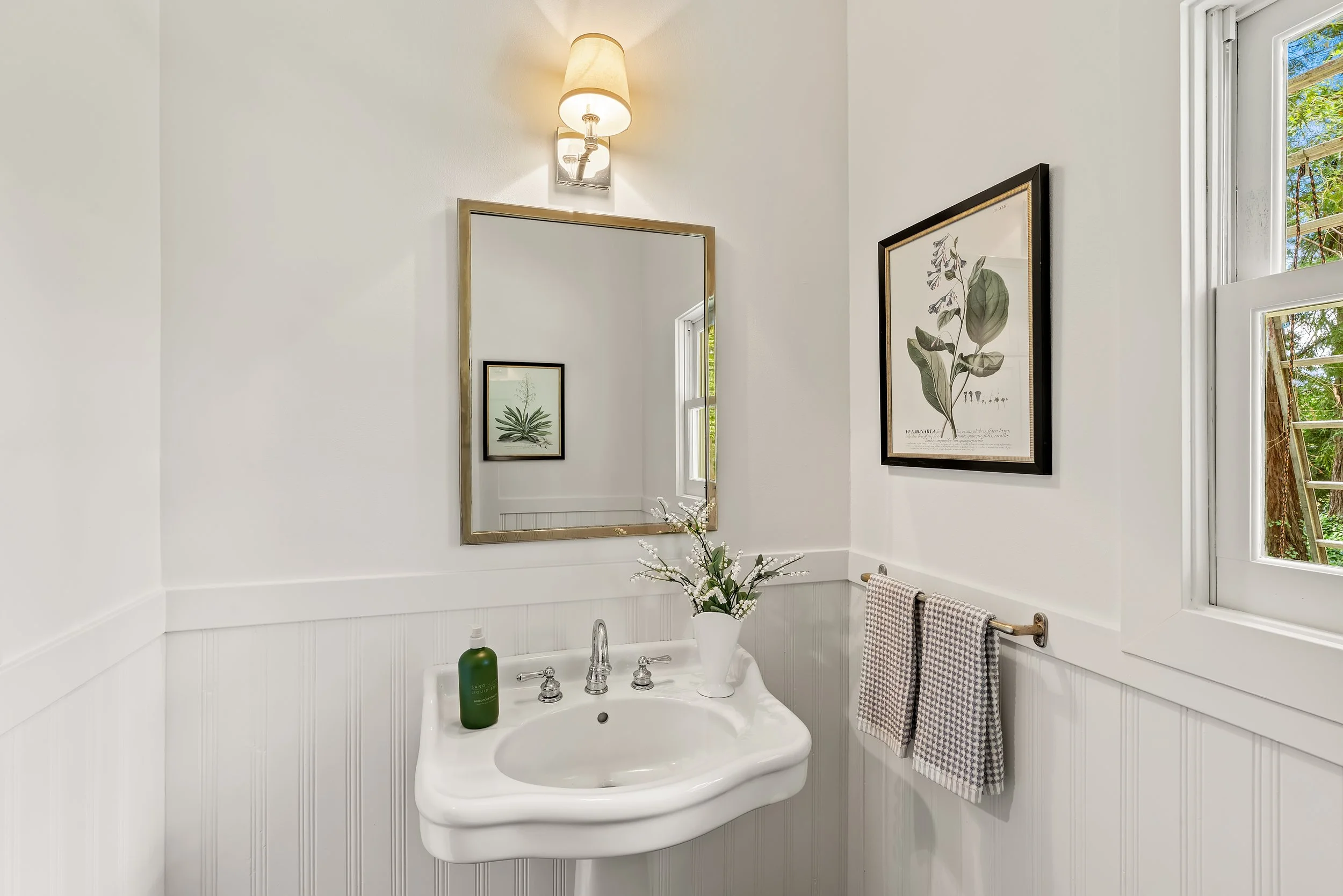 A bathroom with a white vanity, a framed mirror, wall art of plants, a window with a view of trees, a light above the mirror, a vase with flowers, a green soap dispenser, and a checkered towel hanging on a towel bar.