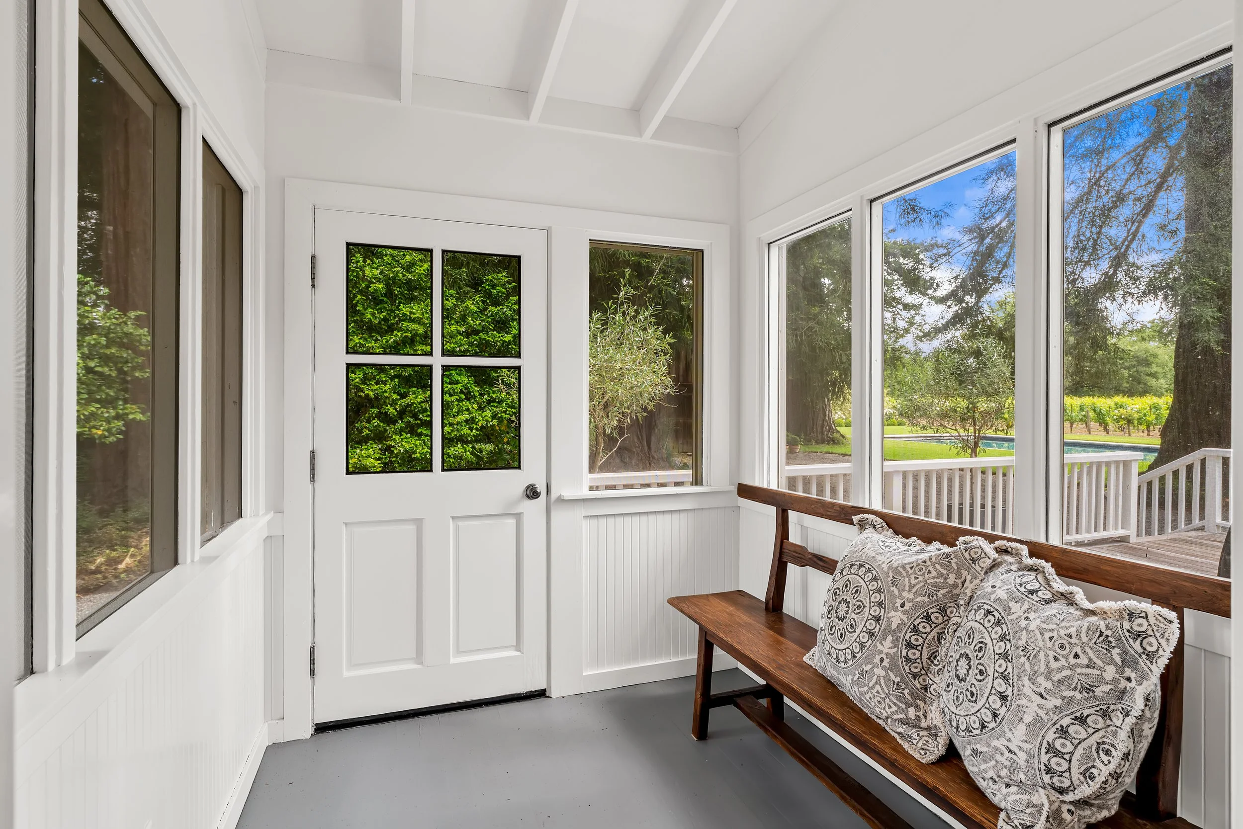 Enclosed porch with white walls and large windows showing greenery outside, wooden bench with patterned cushions, and a white door with glass panes.