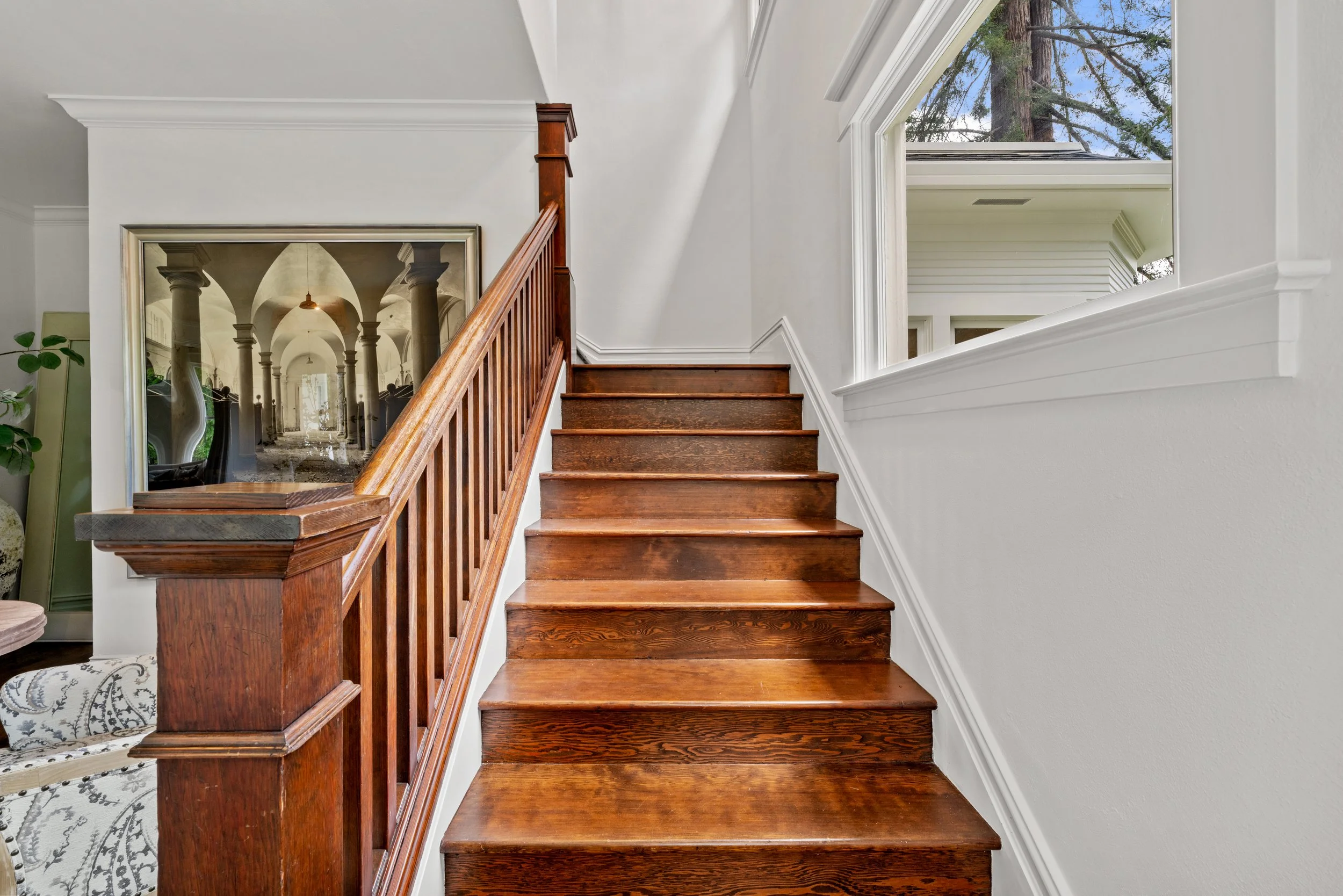 Wooden staircase with white walls and large window revealing a tree outside.