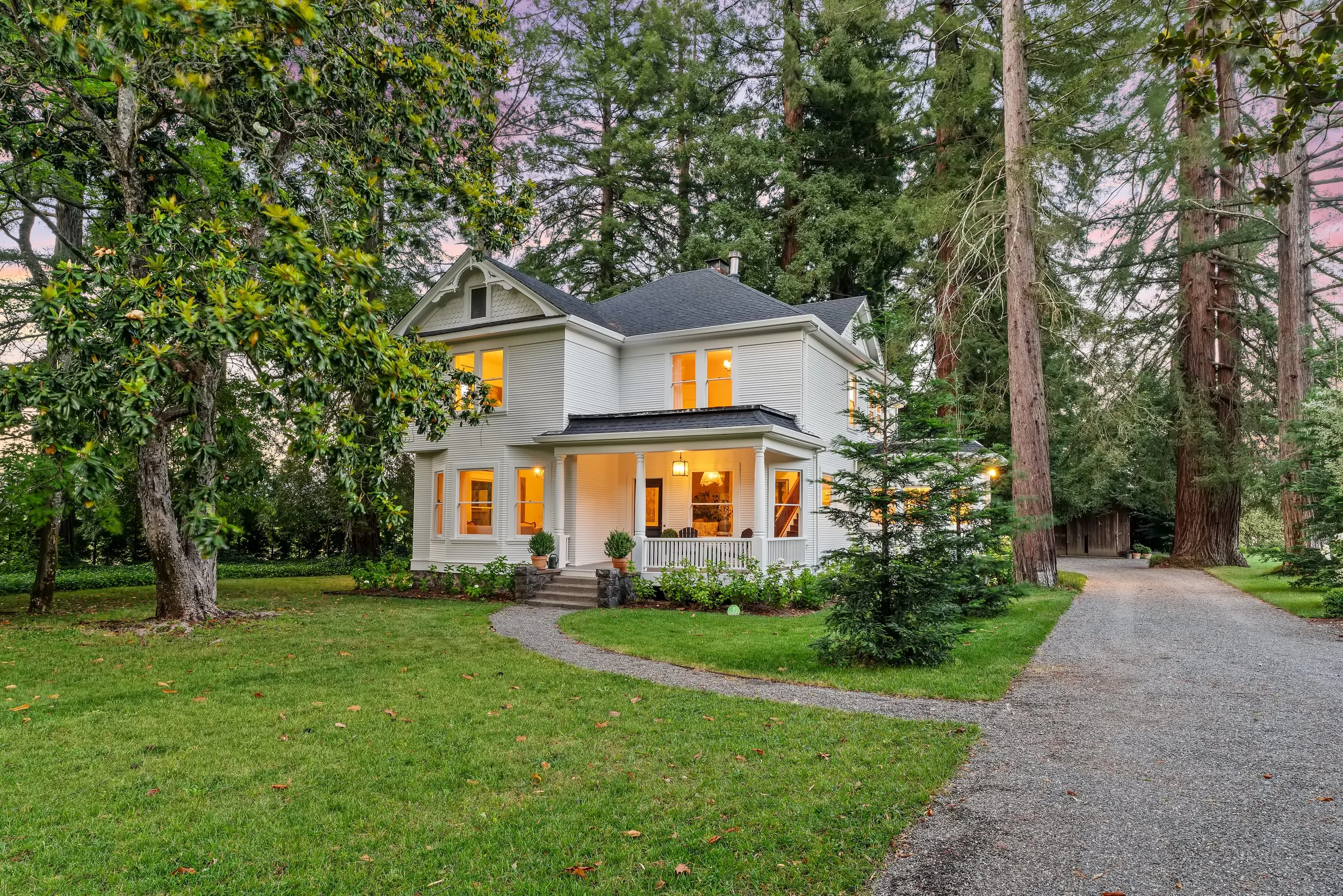 A two-story white house with a porch, surrounded by large trees and green lawn, with a gravel driveway to the right.