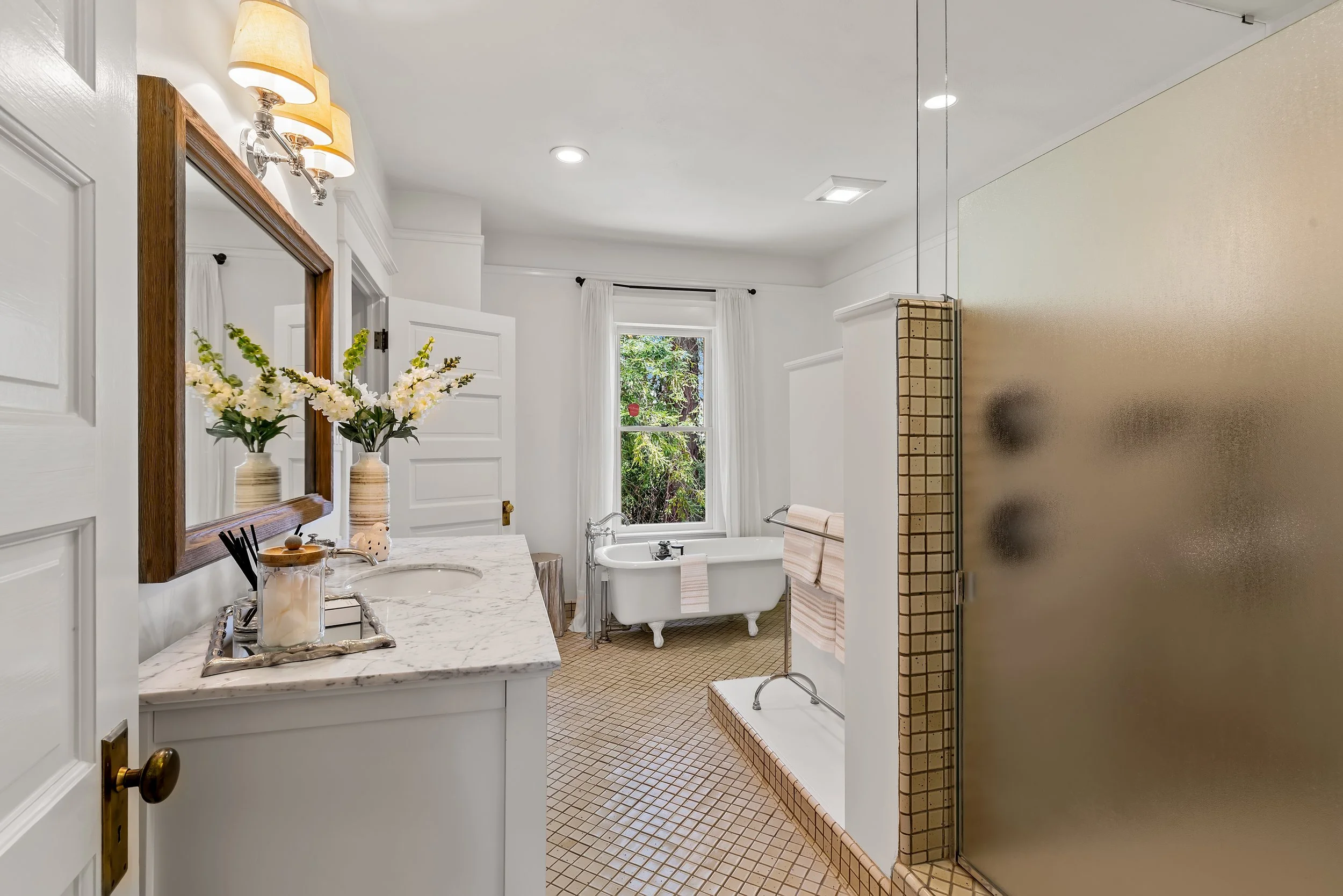 A bright bathroom with white walls and ceiling, featuring a marble countertop with a cow-shaped soap dispenser and small decorative items. A large mirror with a wooden frame hangs above the sink. The bathroom includes two windows with white curtains,