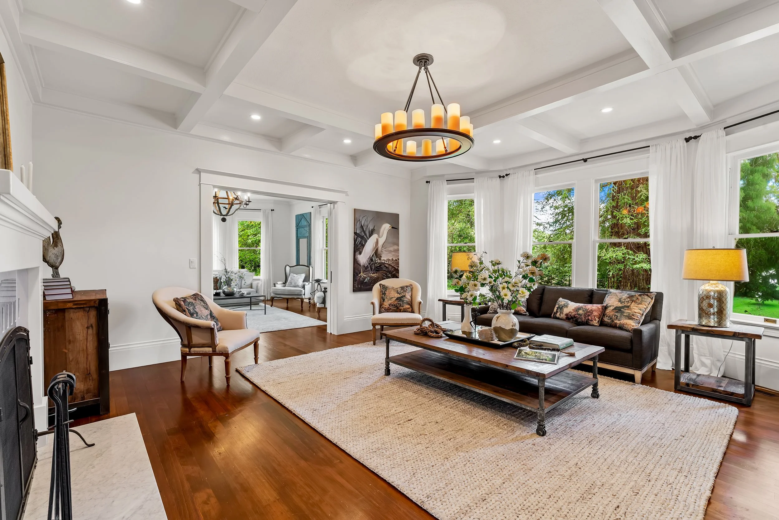 A living room with white walls, large windows with white curtains, a wooden coffee table with decorative items, a beige armchair, a gray sofa with patterned pillows, and a ceiling chandelier with cylindrical lights.