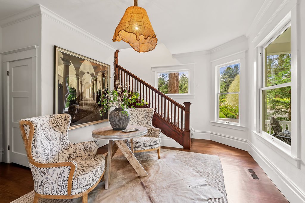 Bright living room with large windows, wooden staircase, patterned armchairs, a round table with a vase of flowers, and a large picture on the wall.