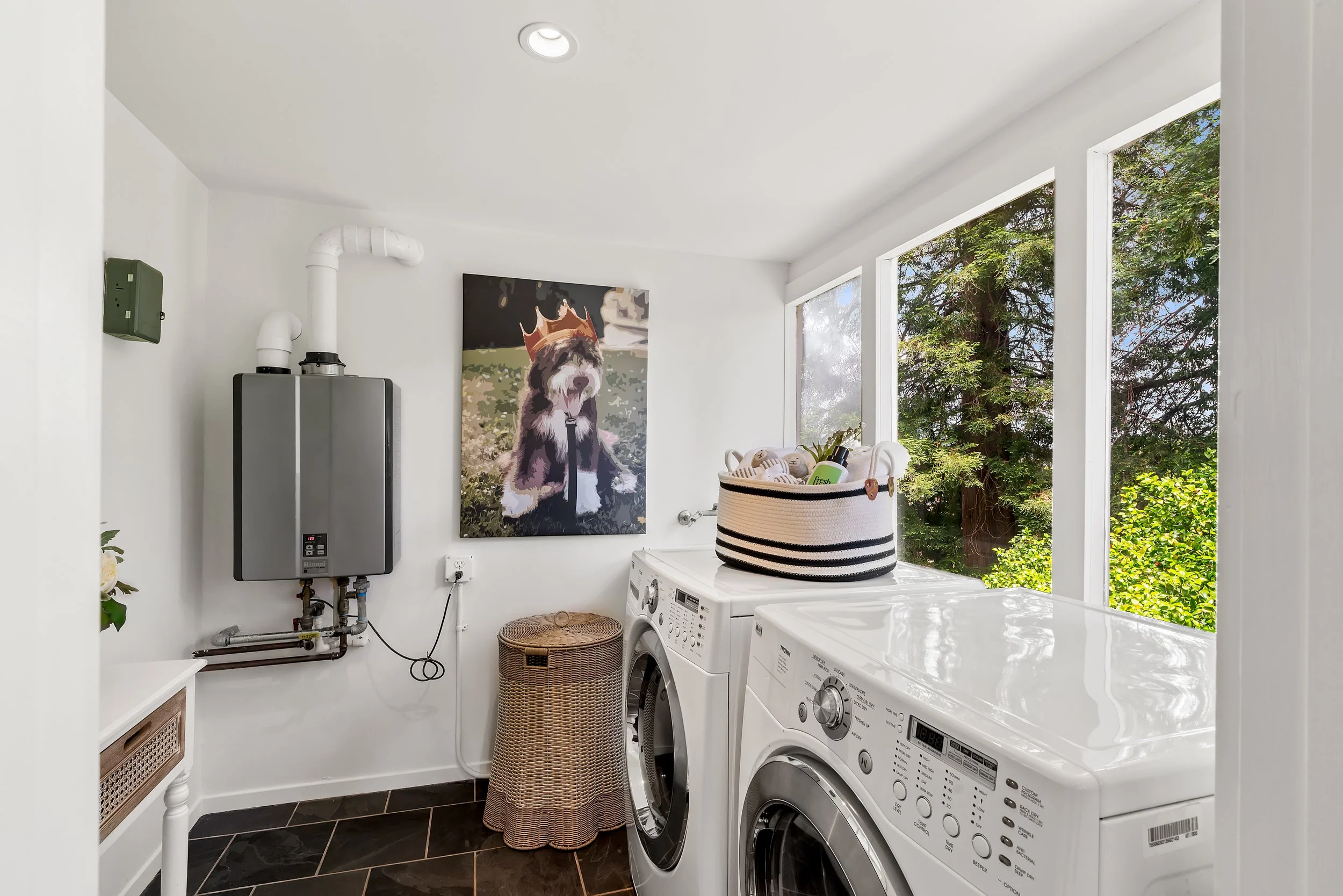 A laundry room with white walls and large windows showing green trees outside. There are two white washing machines, a laundry basket on top of one, and a wall-mounted water heater. A framed painting of a dog wearing a crown is on the wall.