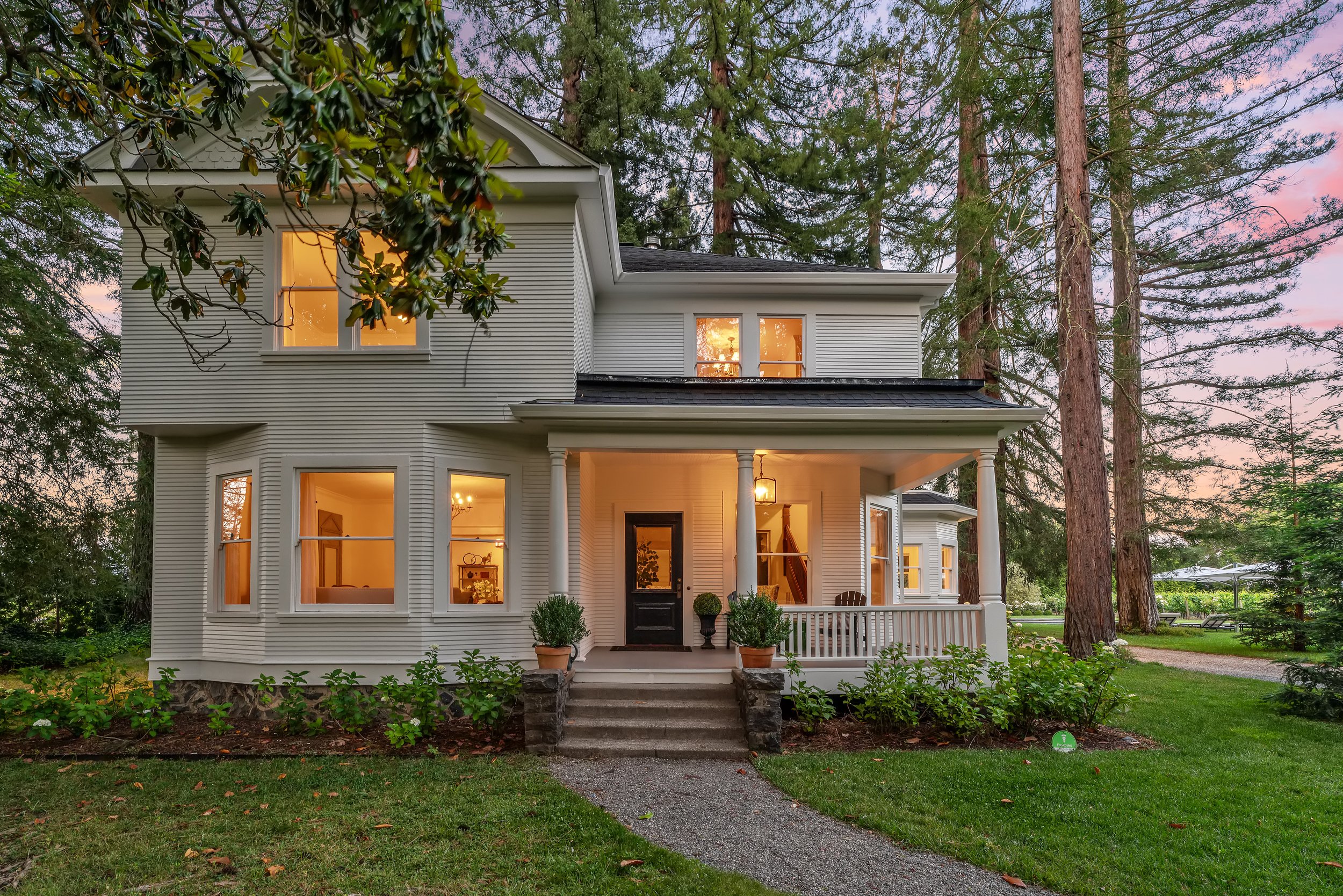A white, two-story house with large windows, front porch, and white columns, surrounded by tall trees and green grass during dusk with warm interior lighting visible.