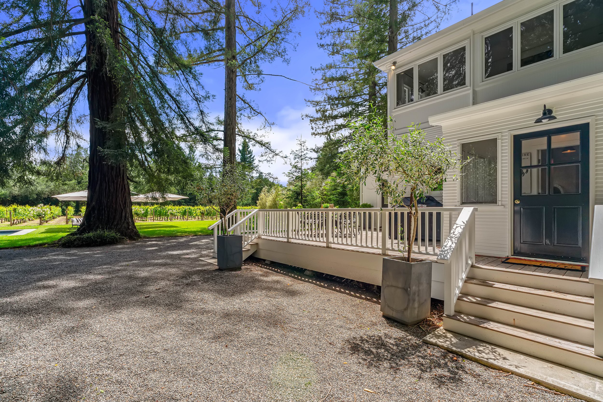 A white two-story house with a black door and large windows on the upper floor, surrounded by greenery and tall trees, with a small deck and stairs leading to the ground.