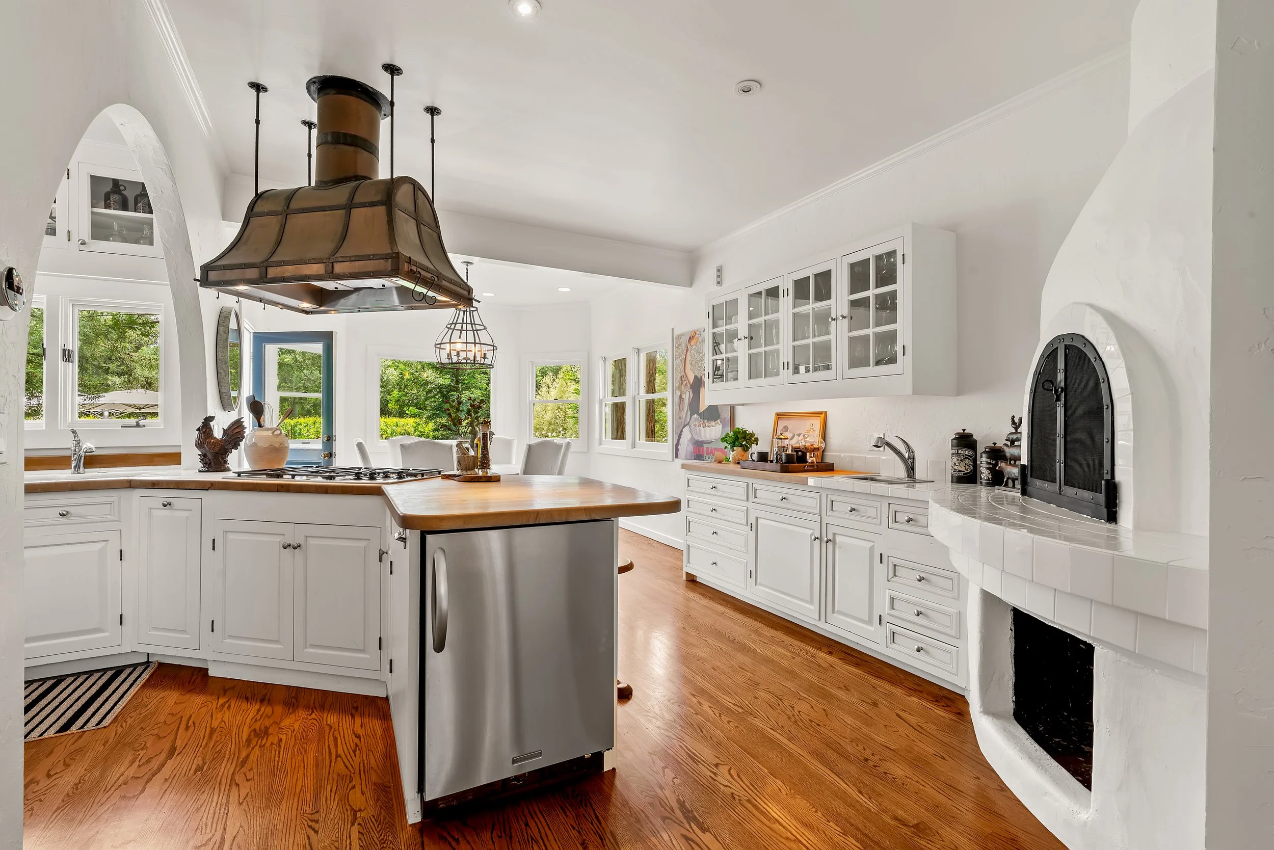 Bright white kitchen with wood floors, white cabinets, and a large central island. Black metal range hood above the stove, glass-fronted cabinets on the wall, and windows showing green trees outside. Kitchen decor includes a rooster statue and vases.