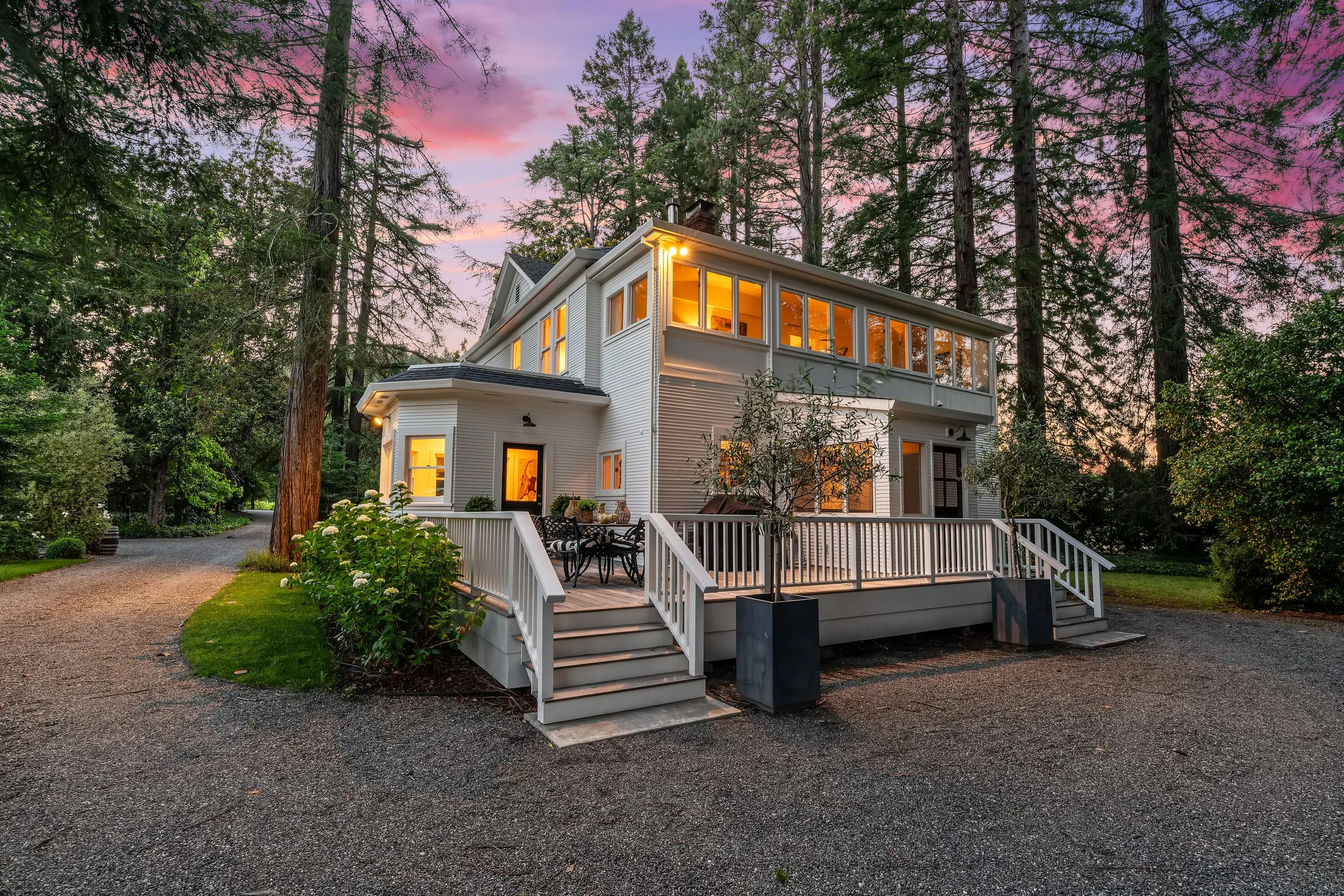 A modern white house surrounded by tall trees, with a porch and outdoor dining area at sunset. The sky is painted with pink and purple hues, and the house's interior lights are on.