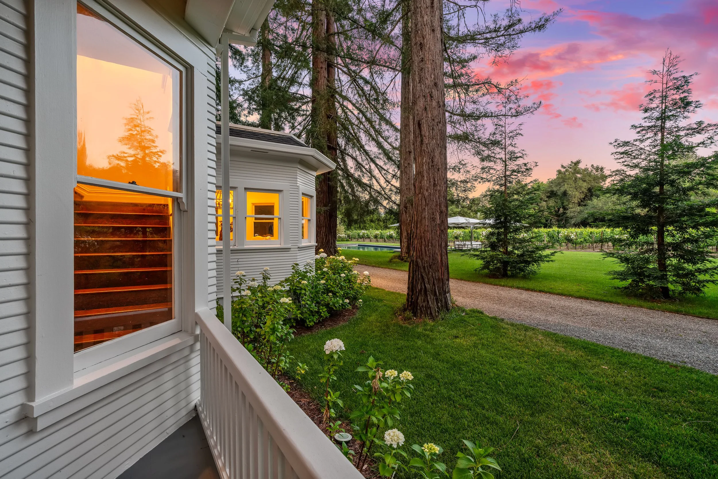 View of a house with white siding and large windows reflecting trees at sunset, surrounded by lush green lawn, tall trees, and a gravel pathway under a colorful sky with pink and purple clouds.