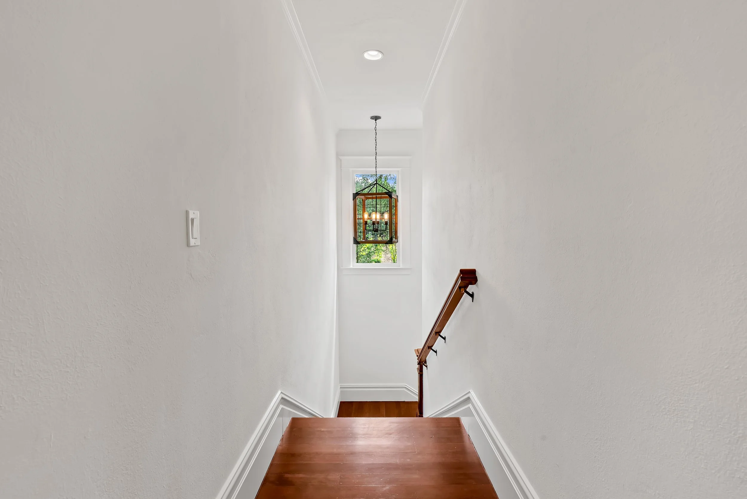 A staircase with wooden steps and a wooden handrail on the right, leading up to a window with a view of green trees, white walls, a chandelier hanging from the ceiling, and a ceiling light.