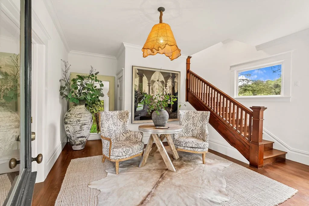 A cozy corner in a living space with two patterned armchairs around a round wooden table, a large vase with green foliage on the table, a tall vase with greenery in the corner, a large framed artwork on the wall, a wooden staircase, a window showing 