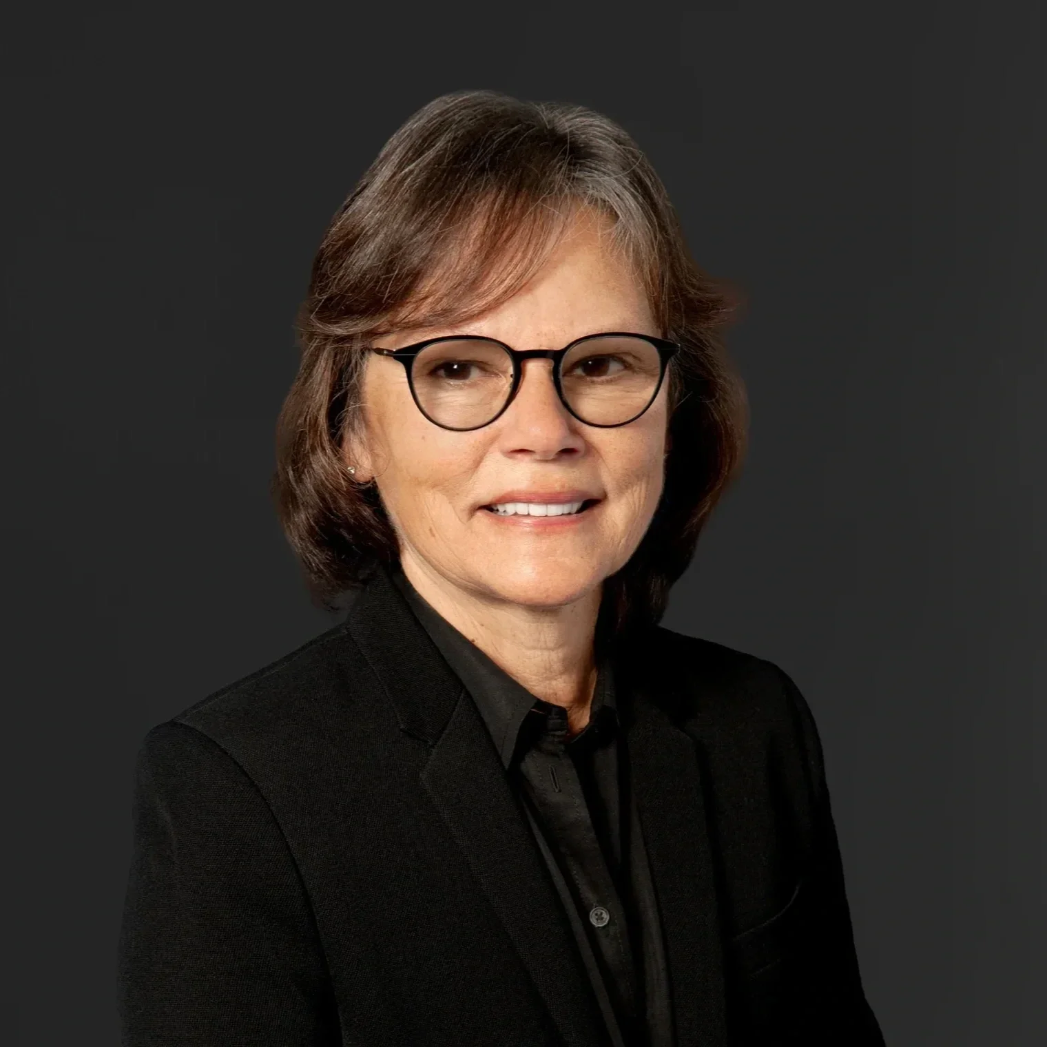 A woman with short brown hair, glasses, and a black blazer, smiling against a dark background.