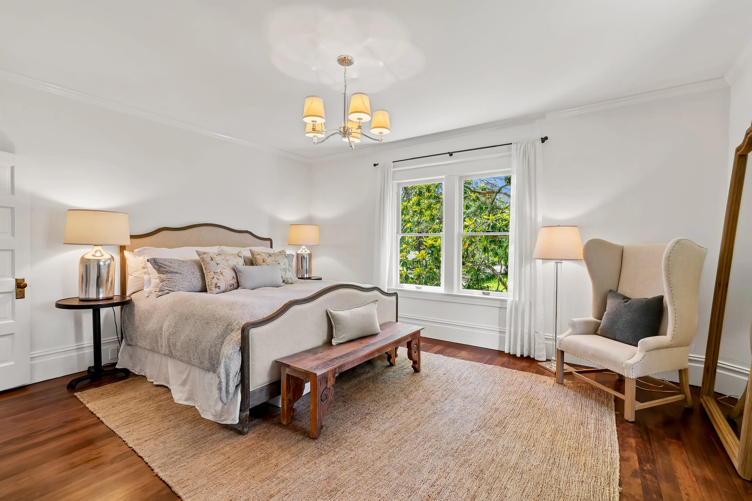 A cozy bedroom with a white bed, two nightstands with lamps, a large window with white curtains, a beige armchair, a wooden bench at the foot of the bed, and hardwood floors with a beige area rug.