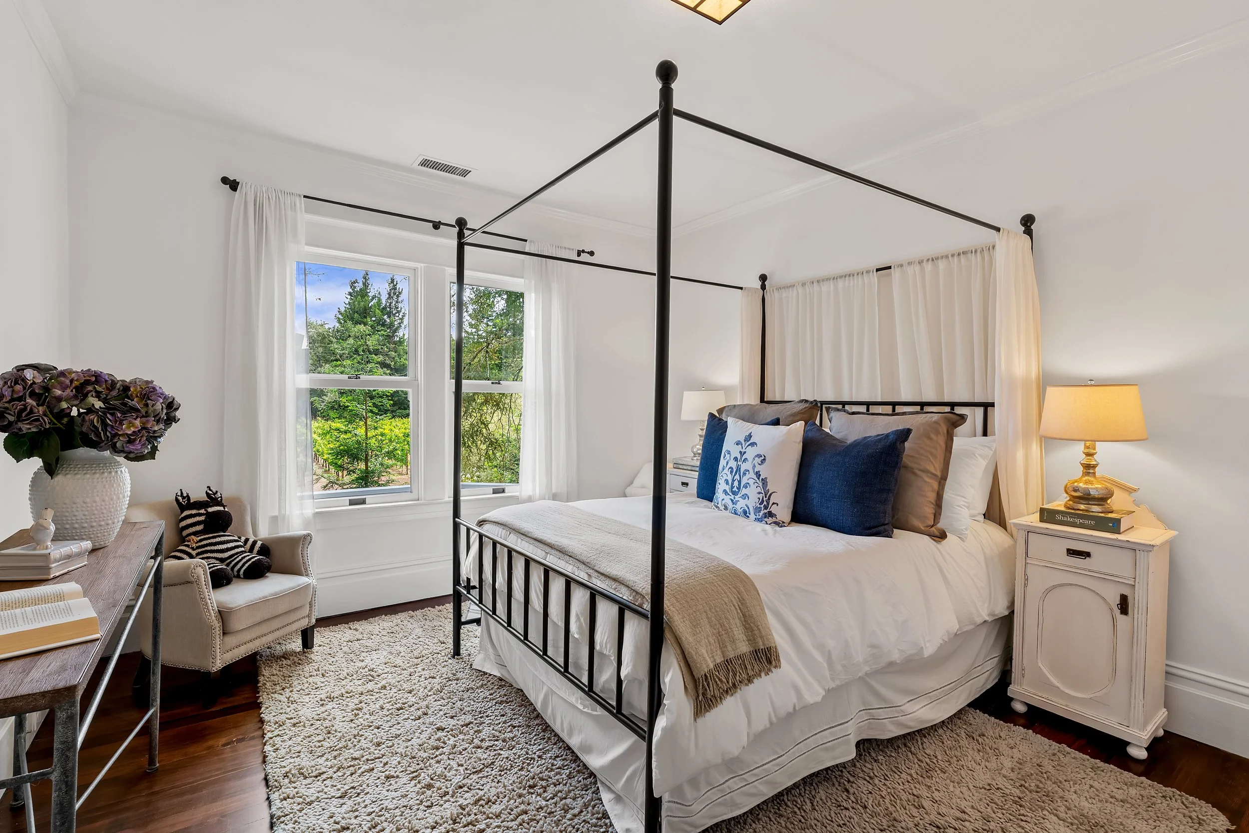 A bedroom with a black metal four-poster bed, white bedding, and decorative pillows. Light-colored curtains hang by the window, and a nightstand with a lamp and books is beside the bed. A vase with purple flowers and a small chair with a stuffed anim