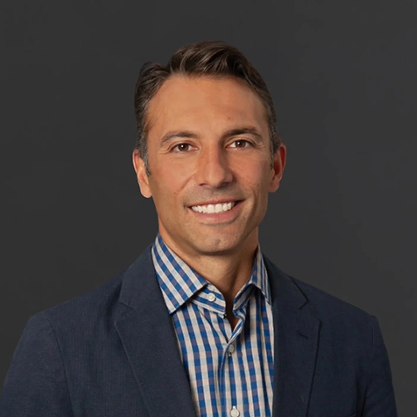 Headshot of a man with short brown hair, wearing a navy blazer and a blue plaid shirt, smiling against a dark grey background.