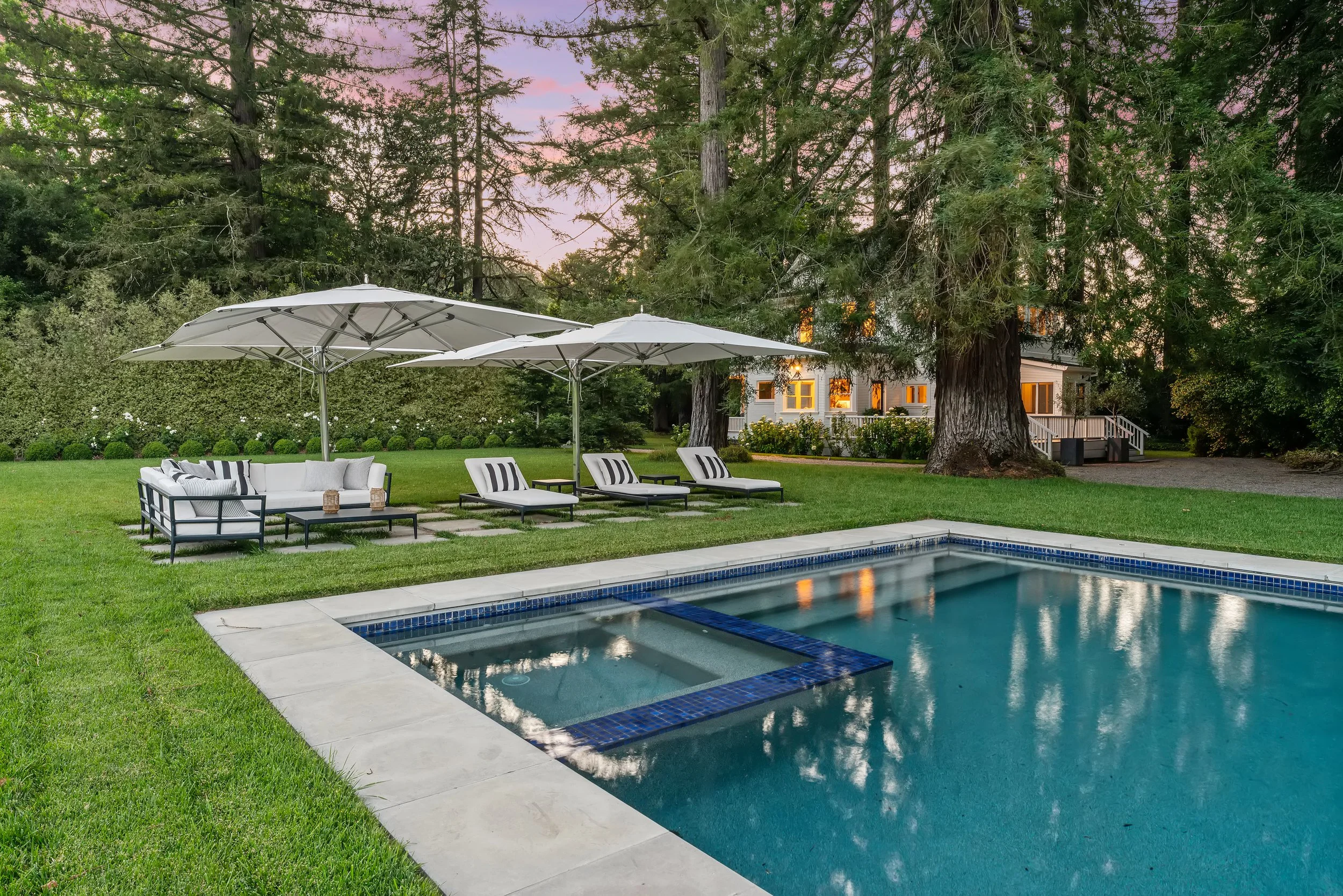 An outdoor swimming pool surrounded by a well-manicured lawn, with lounge chairs and umbrellas on one side, large trees in the background, and a house with large windows and stairs in the distance at sunset.