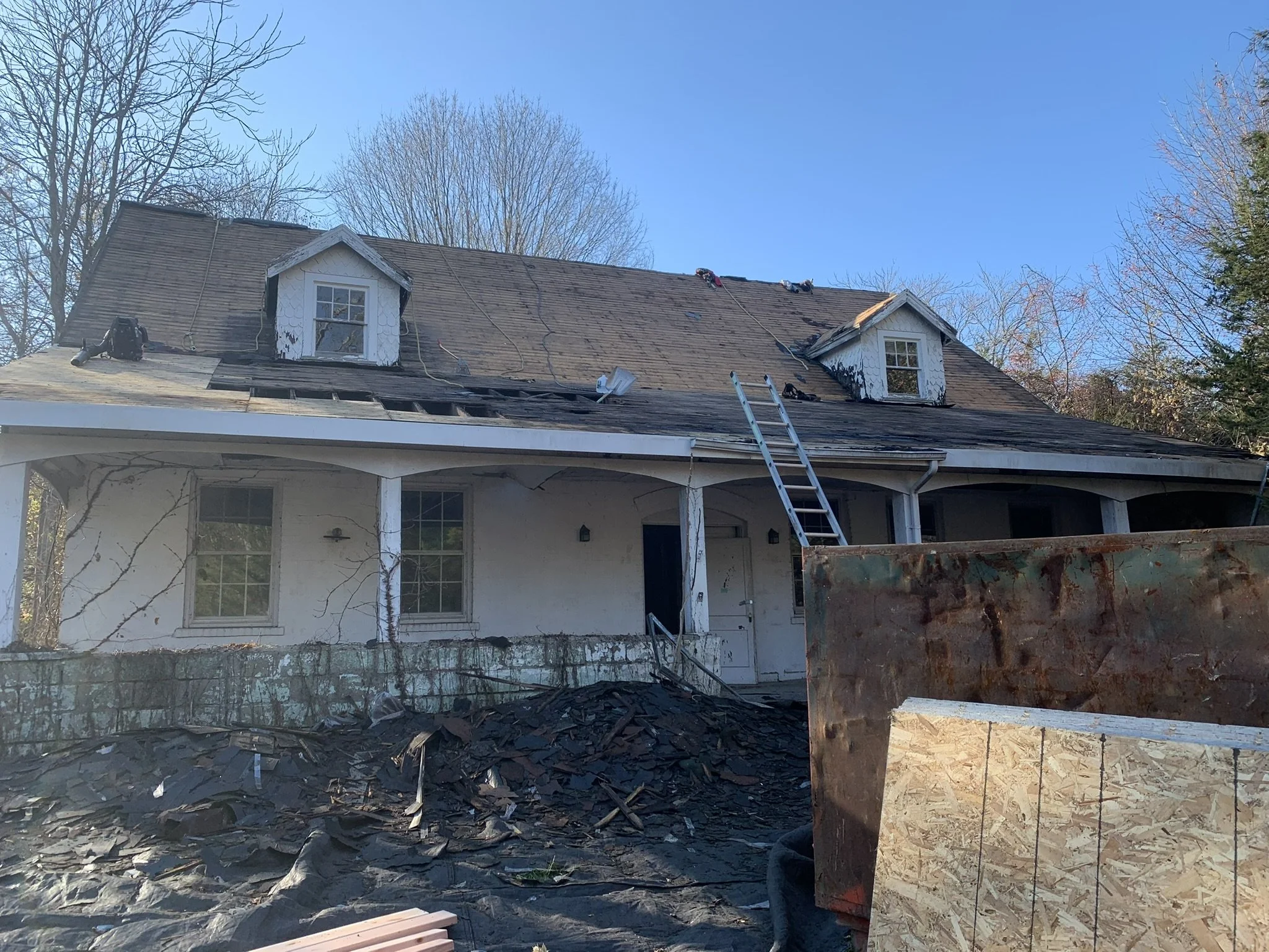 House undergoing roof repair with shingles removed from the roof and roofing tools on top, blue sky, and leafless trees in the background.