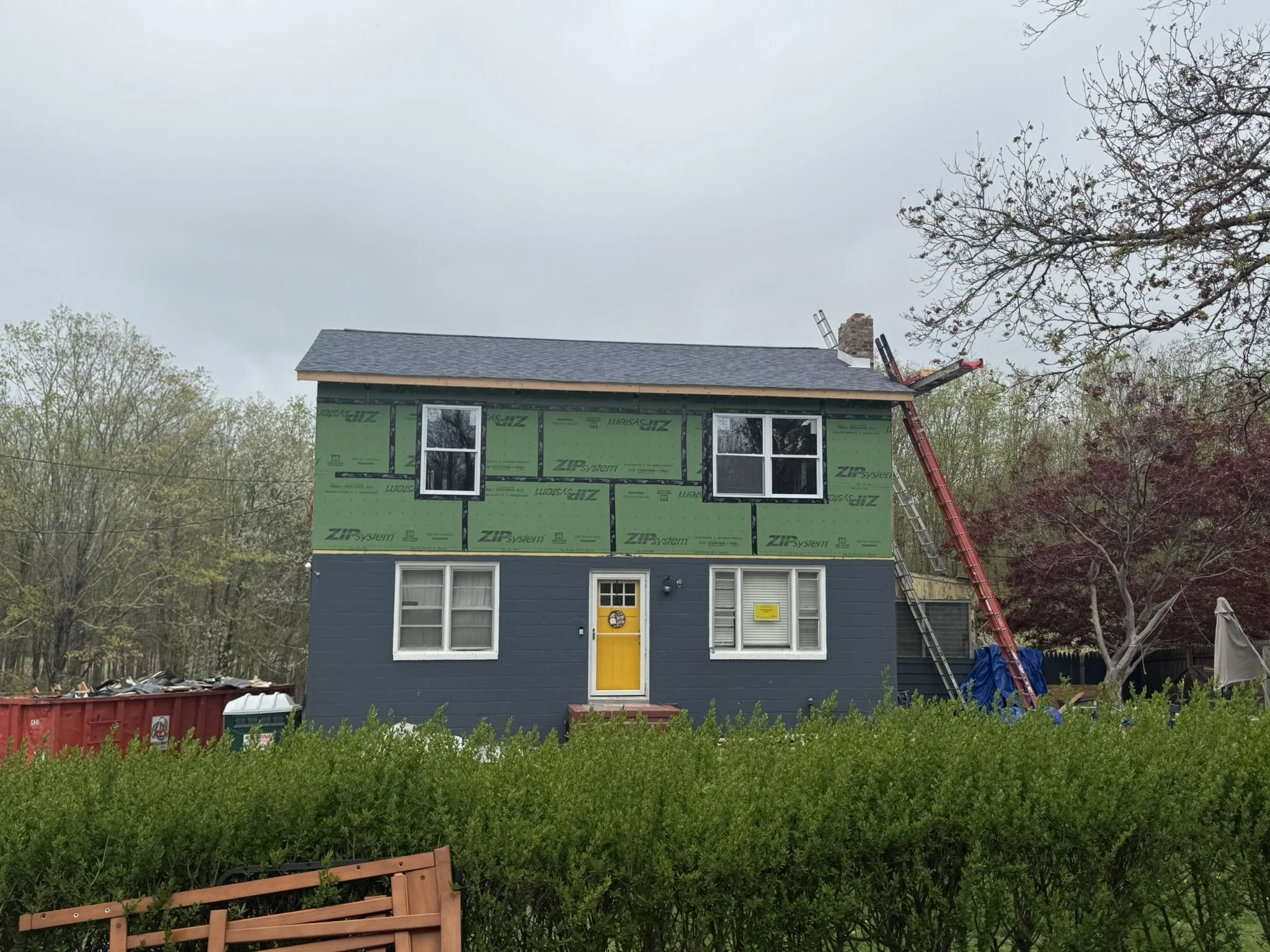 A house under construction with green sheathing on the upper floor, a yellow front door, and a blue lower level. There are windows on both floors, a ladder leaning against the roof, and a chimney. In the foreground, greenery and a wooden bench are visible, with trees and a cloudy sky in the background.