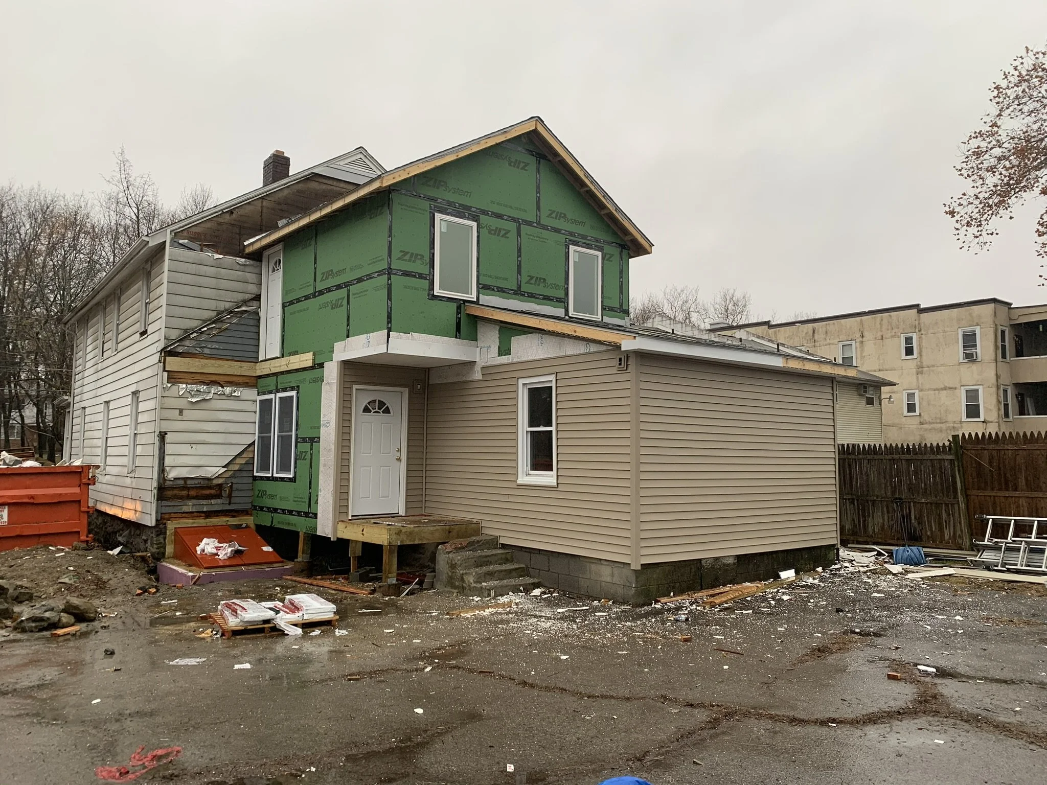 A house under construction with an unfinished upper story, green sheathing, and construction materials scattered on muddy ground in a neighborhood with other residential buildings, leafless trees, and an overcast sky.