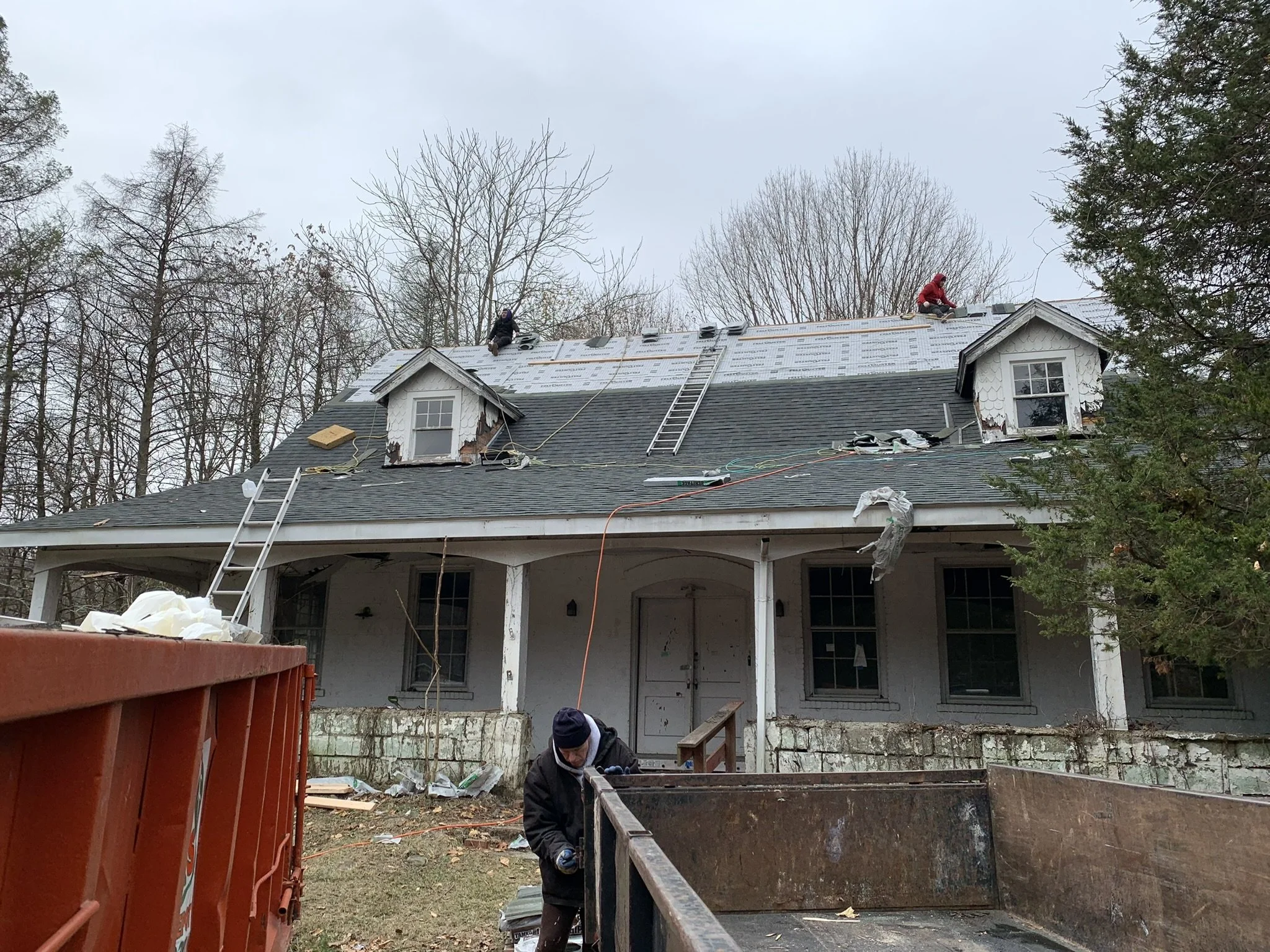 Two workers on the roof of a house installing new roofing shingles, with trees in the background and a large red trash bin in the foreground.