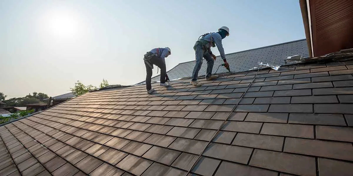 Two workers on a sloped rooftop installing or repairing roof shingles, with tools and safety gear.