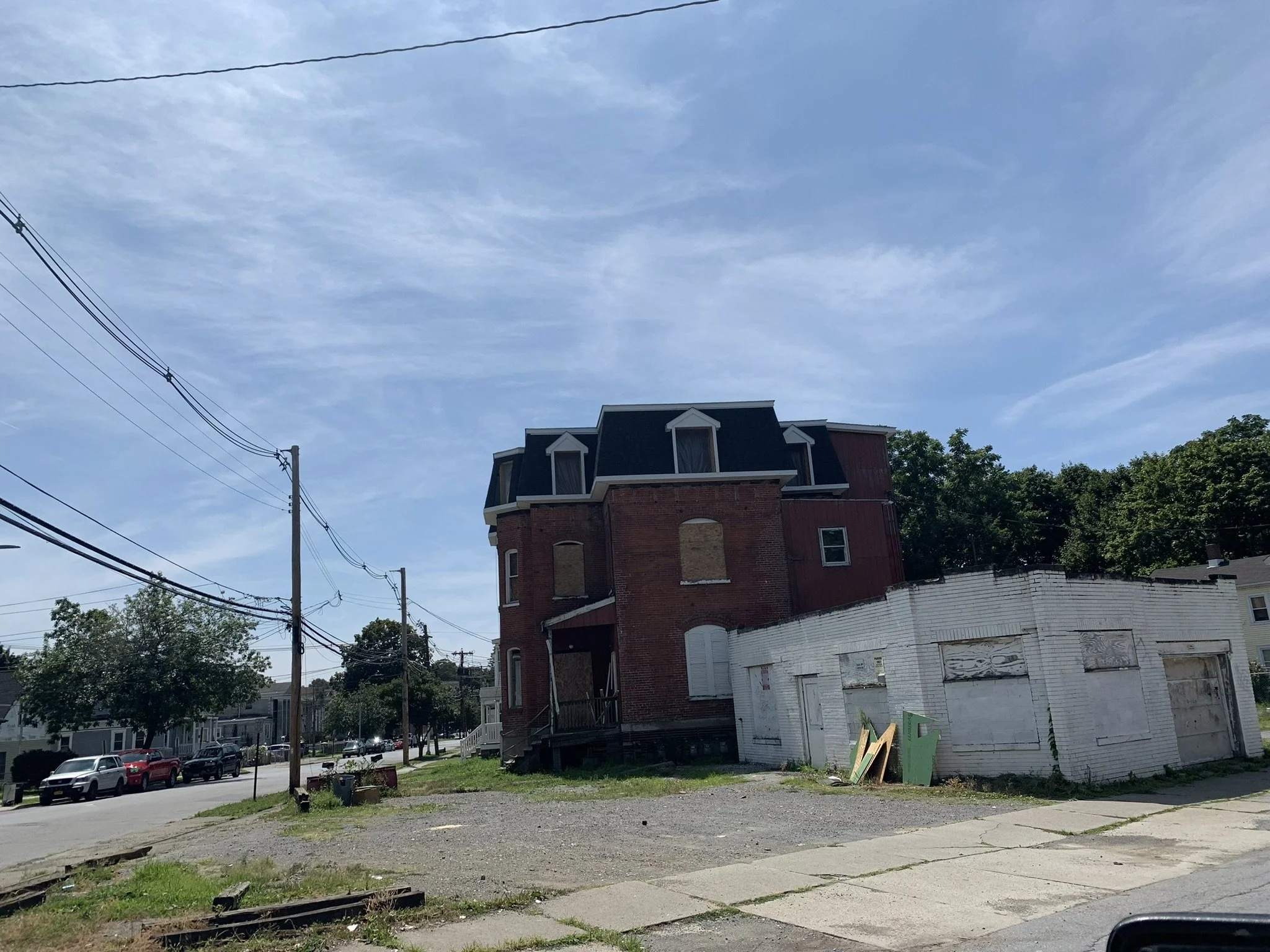 A residential street scene with a multi-story brick house at the corner, featuring a blue roof and white trim. Parked cars line the street, and there are trees and utility poles with wires overhead. A white building with boarded windows is in the foreground on the right, with some lawn and sidewalk in front.