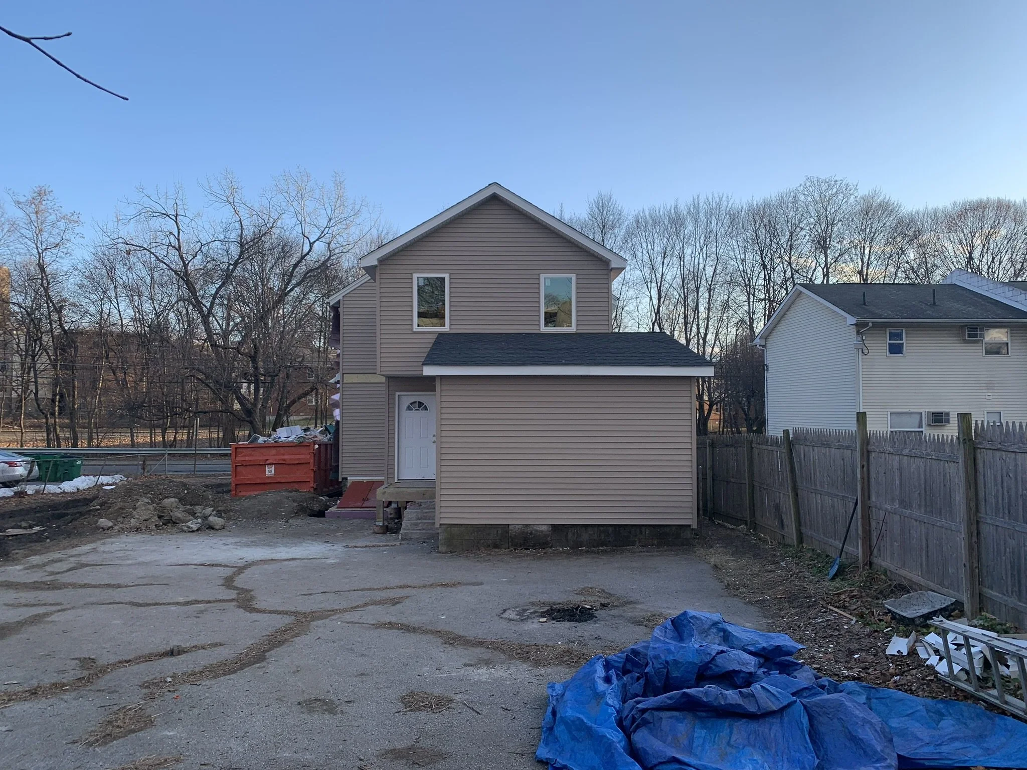 Backyard with a small newly built house, construction debris, and tools scattered around. Bare trees and neighboring houses are visible in the background under a clear blue sky.