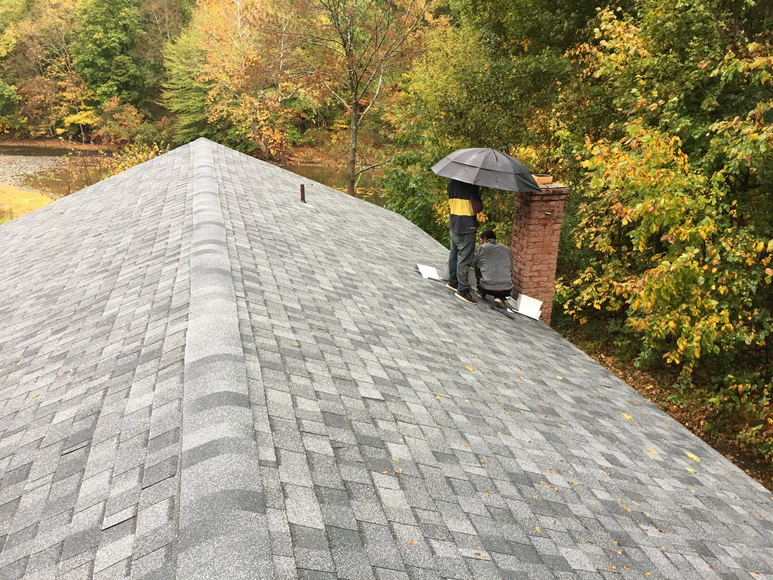 Two people on a rooftop with gray shingles, one holding an umbrella, surrounded by autumn trees and a river in the background.