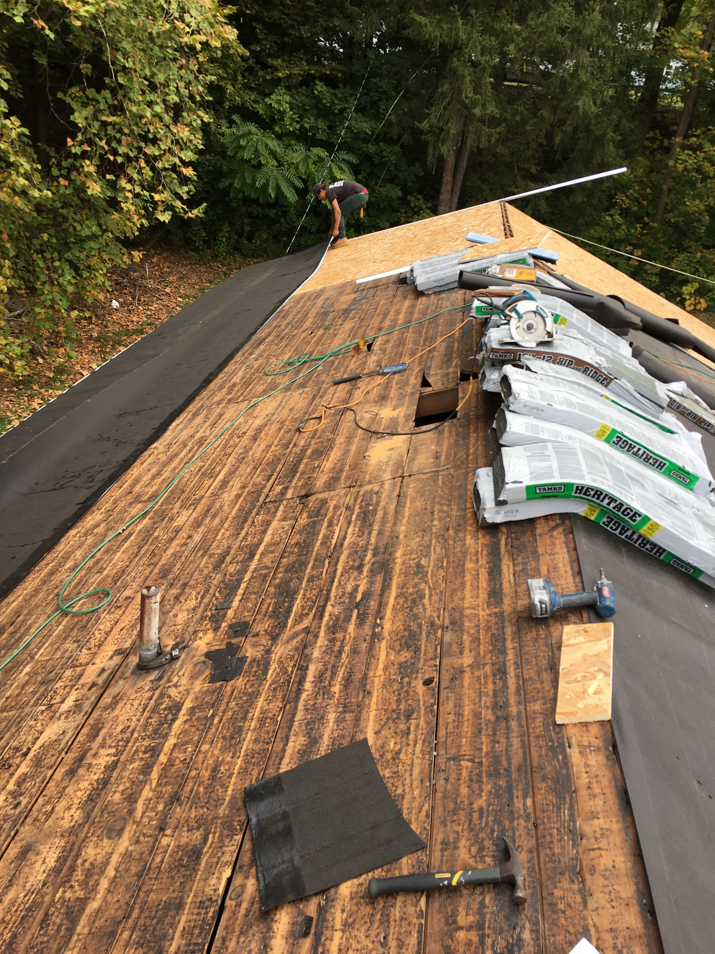 A person working on a roof, installing or repairing underlayment with roofing tools and materials scattered on the wooden roof surface surrounded by trees.