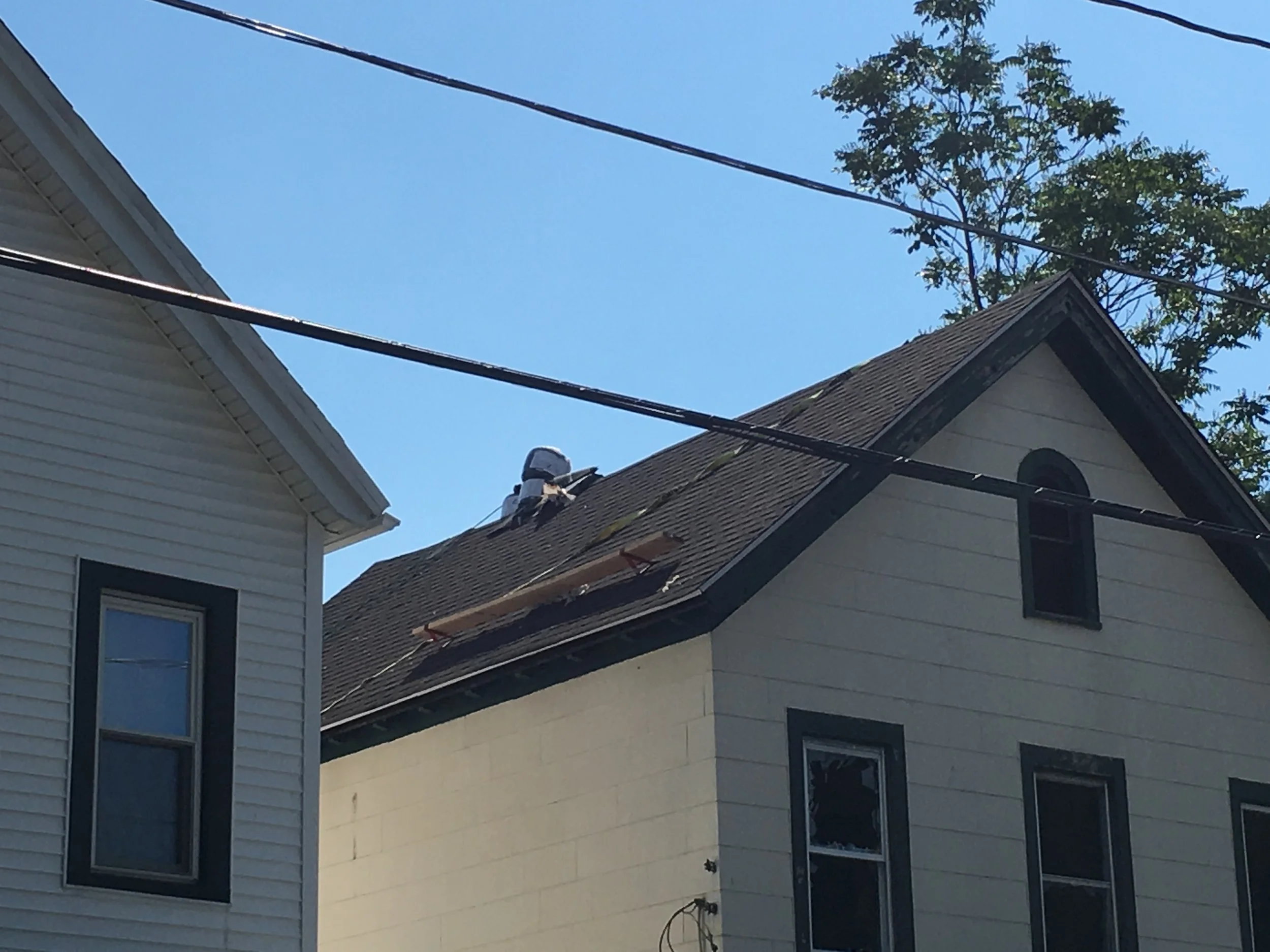 A person working on a roof of a house with shingles, with trees and a blue sky in the background.