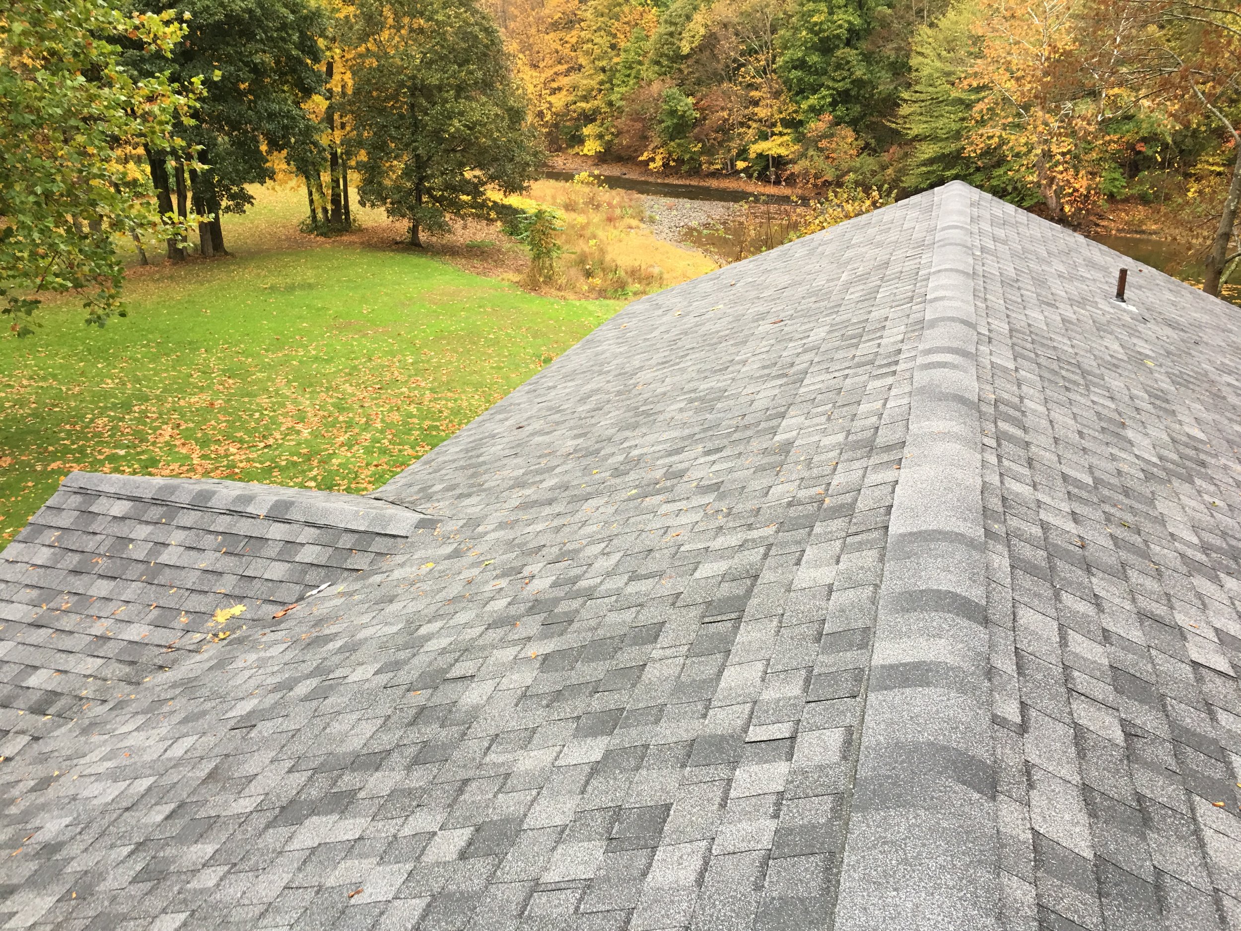 View from a rooftop showing gray asphalt shingles, green grass, deciduous trees with yellow and green leaves, and a small river or creek in the background during fall.