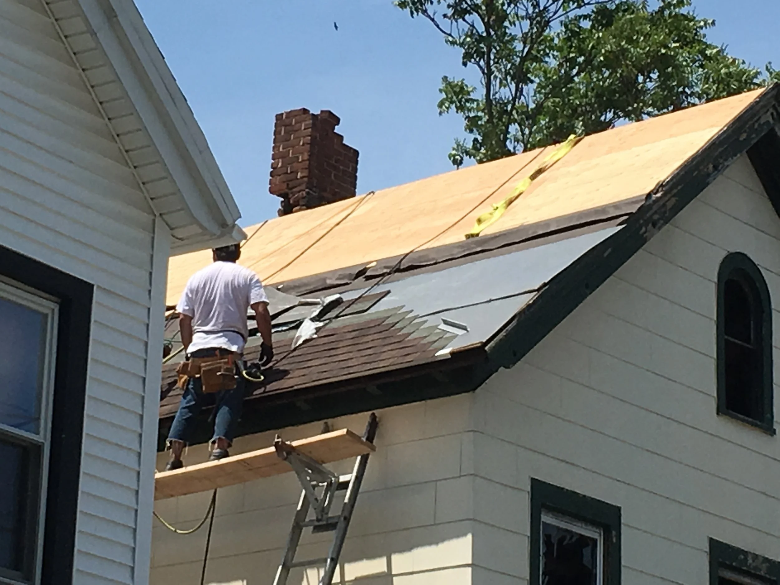 A person working on roofing of a house, standing on a scaffolding platform, with a partially installed roof and a brick chimney visible, under a clear blue sky with trees.